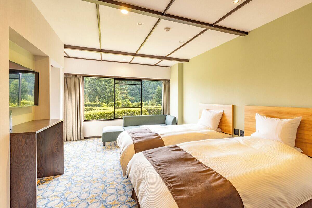 Ein Hotelzimmer mit zwei Betten, einem Schreibtisch, einem Fenster mit Blick auf Grünflächen und einem gemusterten Boden.