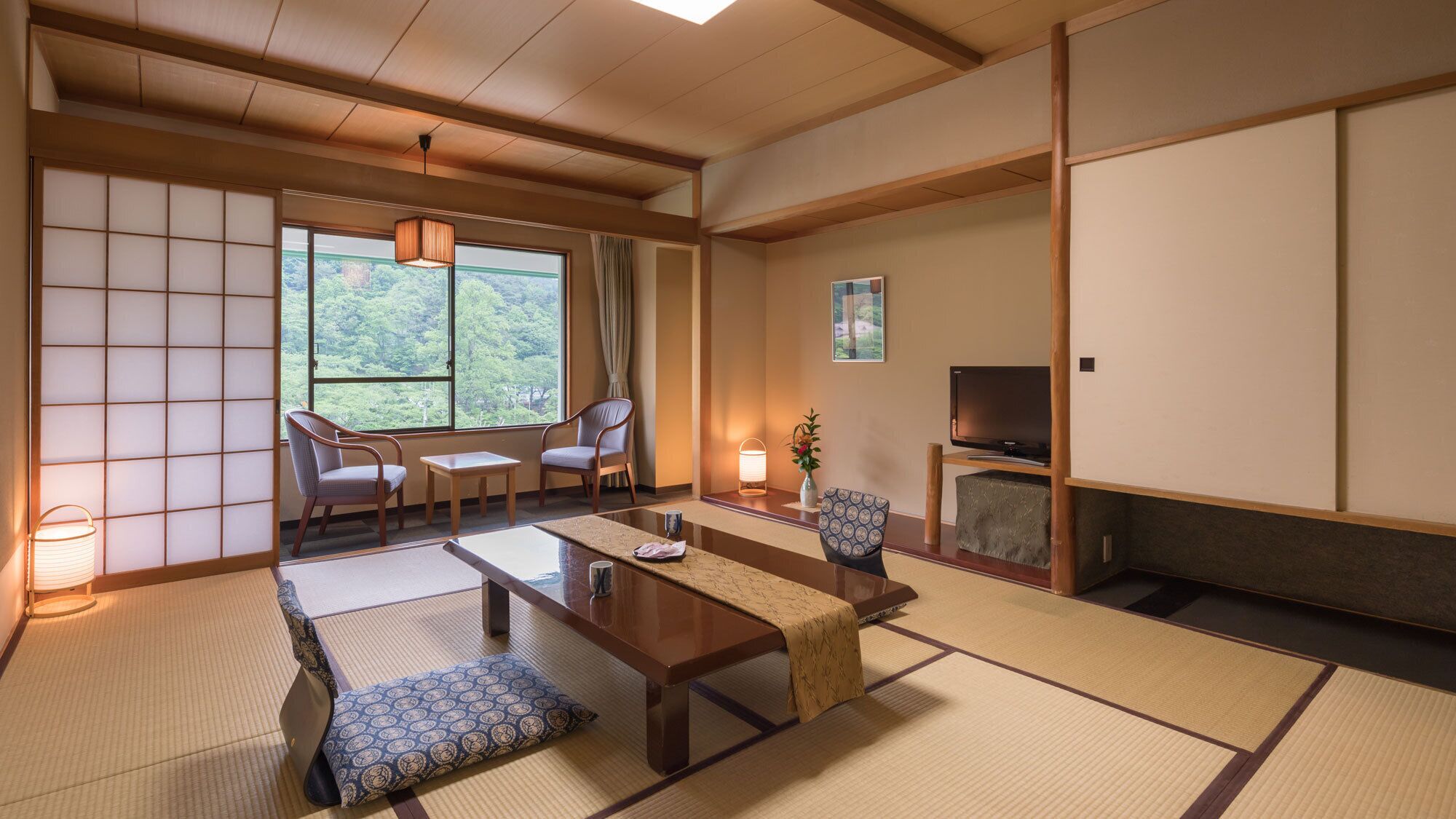 A traditional Japanese room with tatami flooring, a low table, two chairs, a television, and a view of greenery outside.