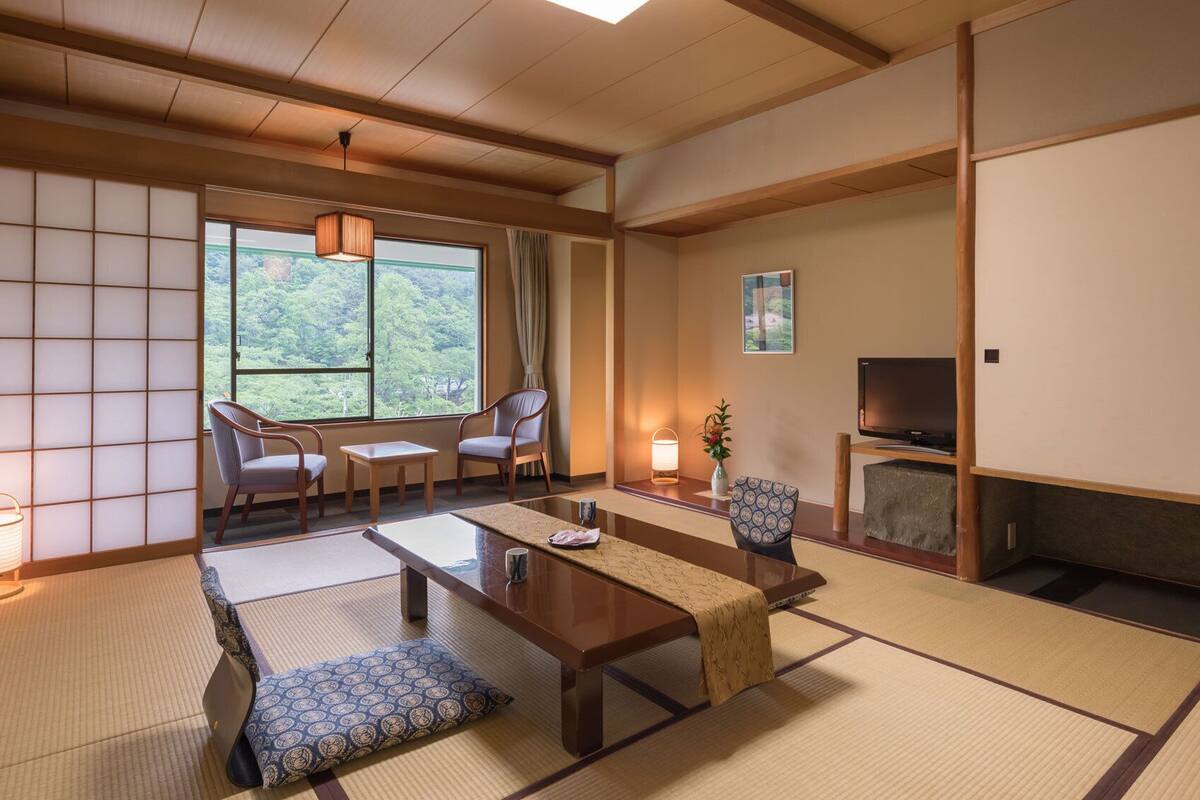 Ein traditioneller japanischer Raum mit Tatami-Boden, einem niedrigen Tisch, zwei Stühlen, einem Fernseher und Blick ins Grüne.