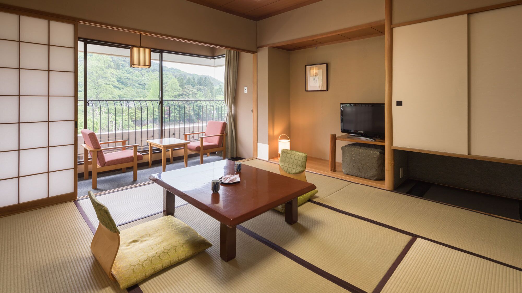 A traditional Japanese room with tatami flooring, a low table, chairs, a television, and a view of greenery outside.