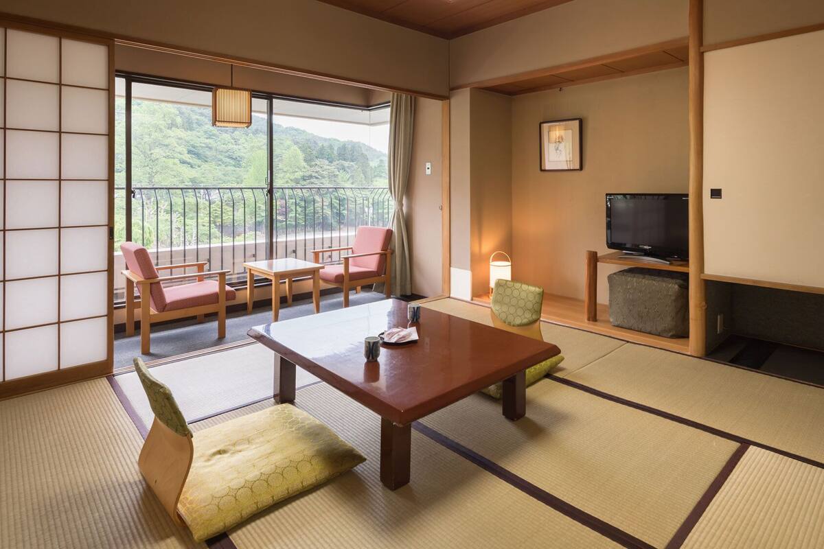 Ein traditioneller japanischer Raum mit Tatami-Boden, einem niedrigen Tisch, Stühlen, einem Fernseher und Blick auf Grünflächen außerhalb.