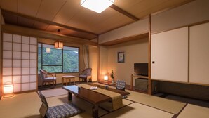 Een traditionele Japanse kamer met tatami vloerbedekking, een lage tafel, twee stoelen, een televisie en een groot raam met uitzicht op groen.