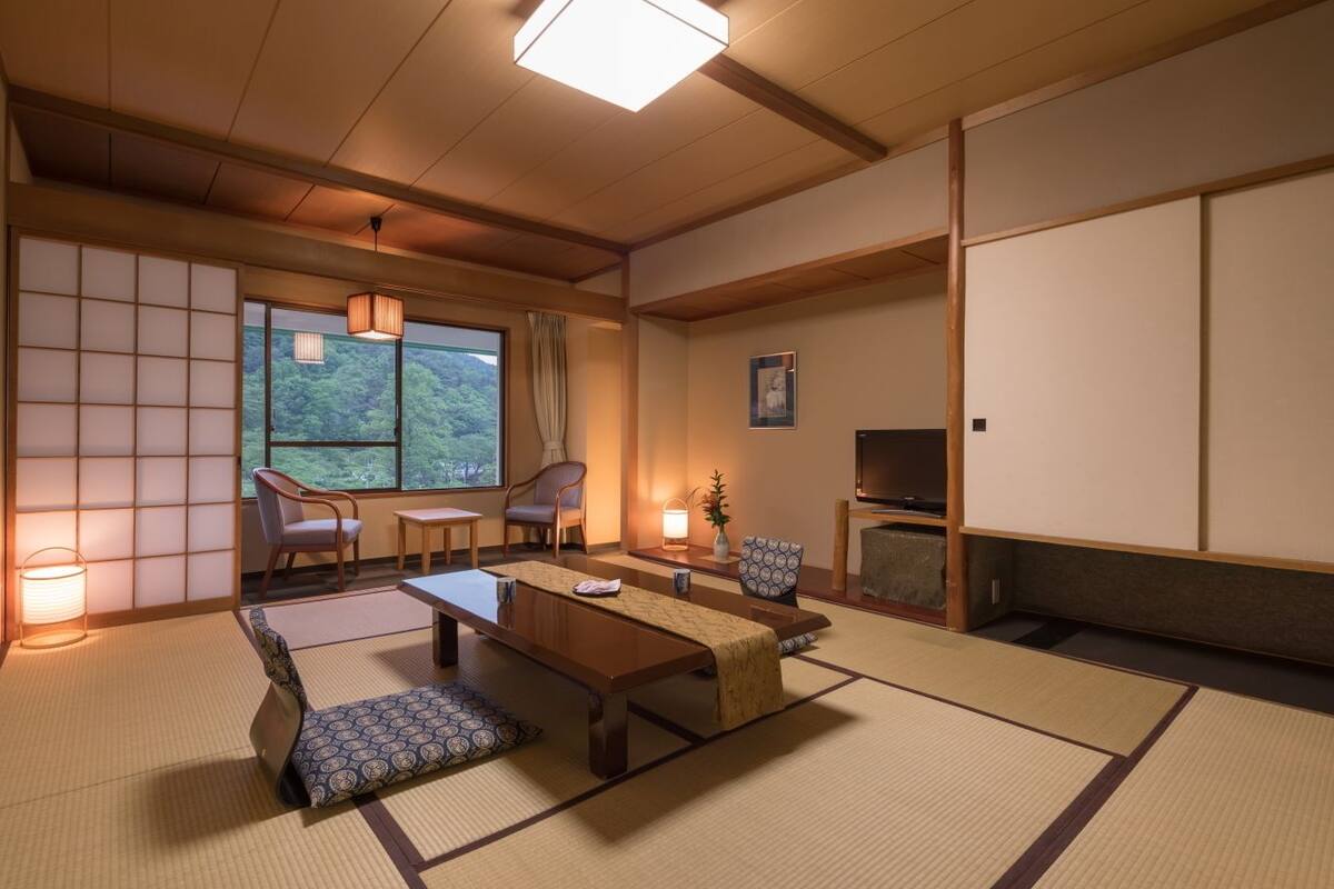 Ein traditioneller japanischer Raum mit Tatami-Bodenbelag, einem niedrigen Tisch, zwei Stühlen, einem Fernseher und einem großen Fenster mit Blick auf Grünflächen.