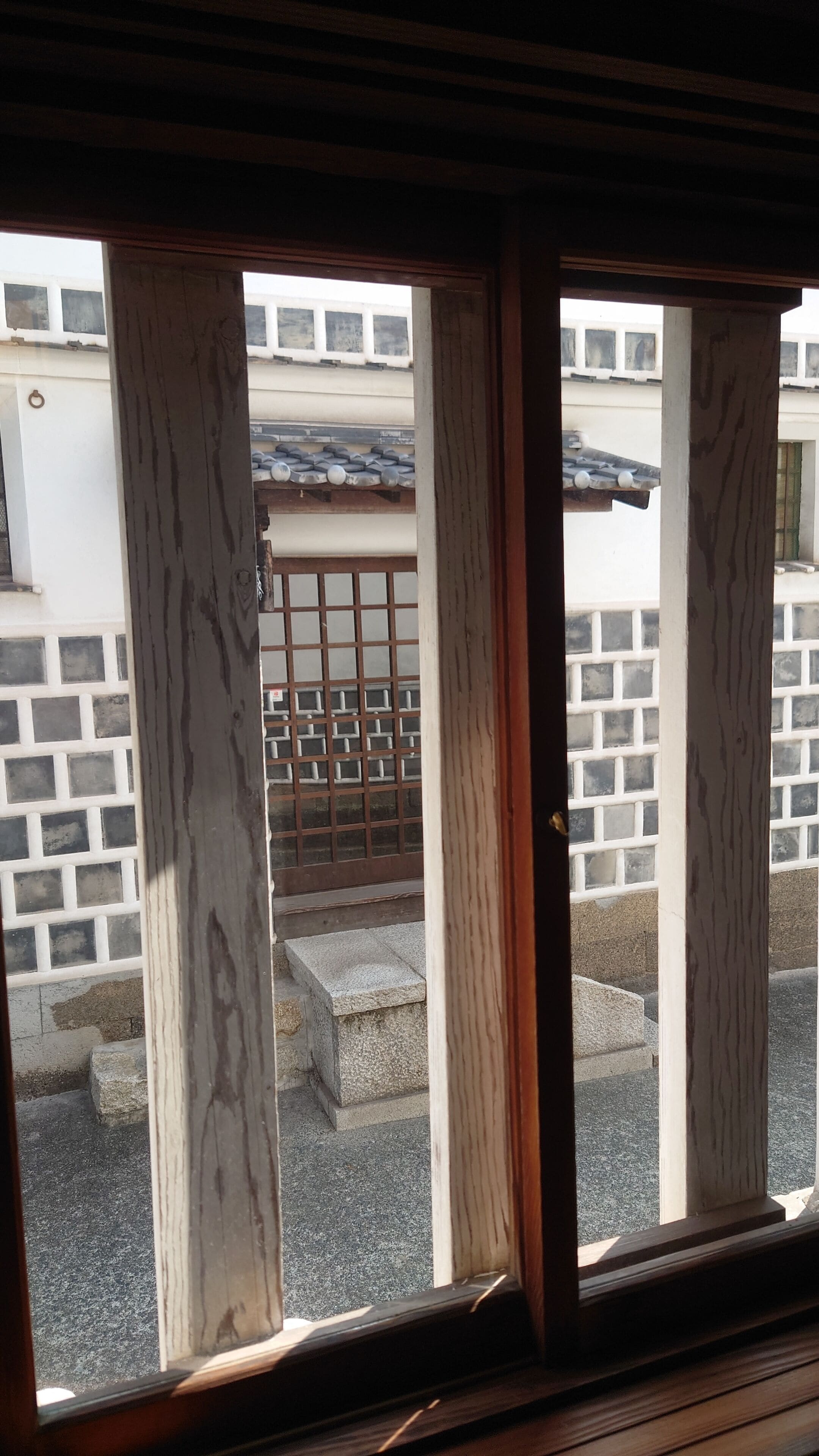 Vista a través de ventanas de madera que muestran un edificio con tejado de baldosas y una puerta de metal.