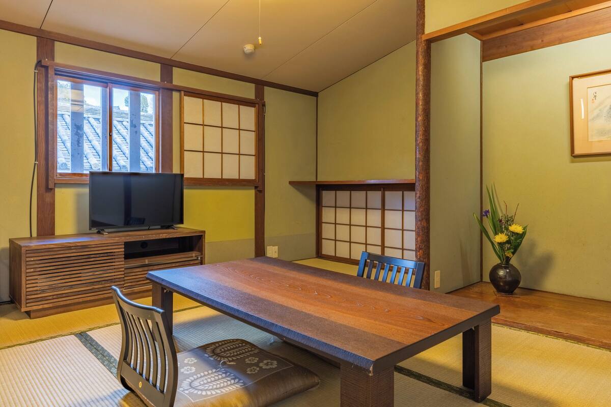 Une pièce de style japonais traditionnel, avec un sol recouvert de tatamis, une basse table en bois, un téléviseur posé sur un meuble en bois et un vase rempli de fleurs.