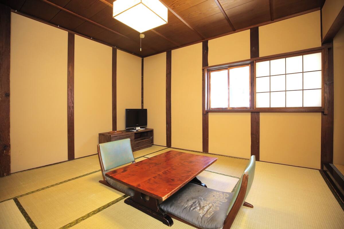Une pièce avec un sol recouvert de tatamis, une petite table basse et des coussins, un téléviseur posé sur un meuble en bois, et une fenêtre à carreaux.