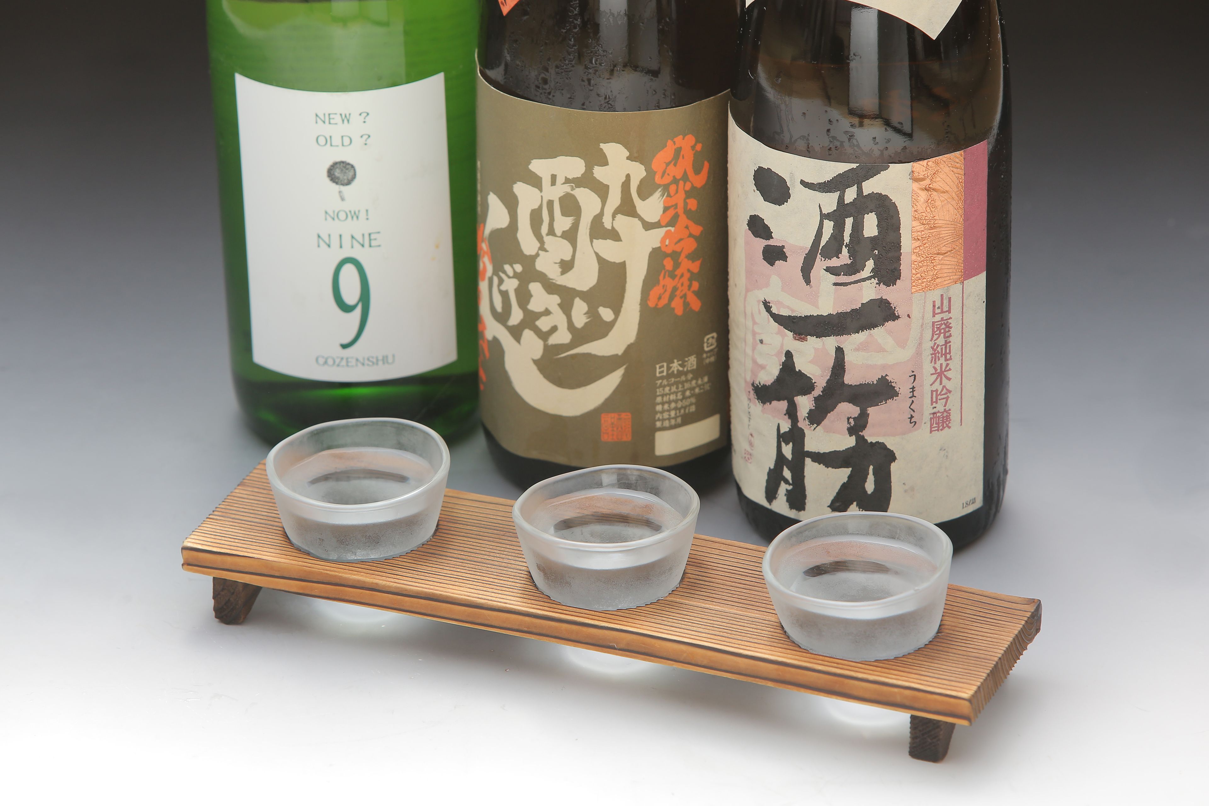 Three bottles of sake with labels, placed on a wooden stand with three small bowls in front.