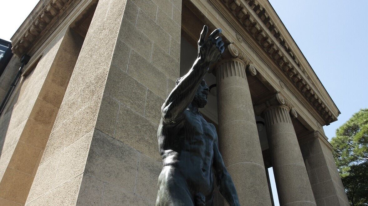 A statue of a man with raised arms in front of a classical building with large columns.