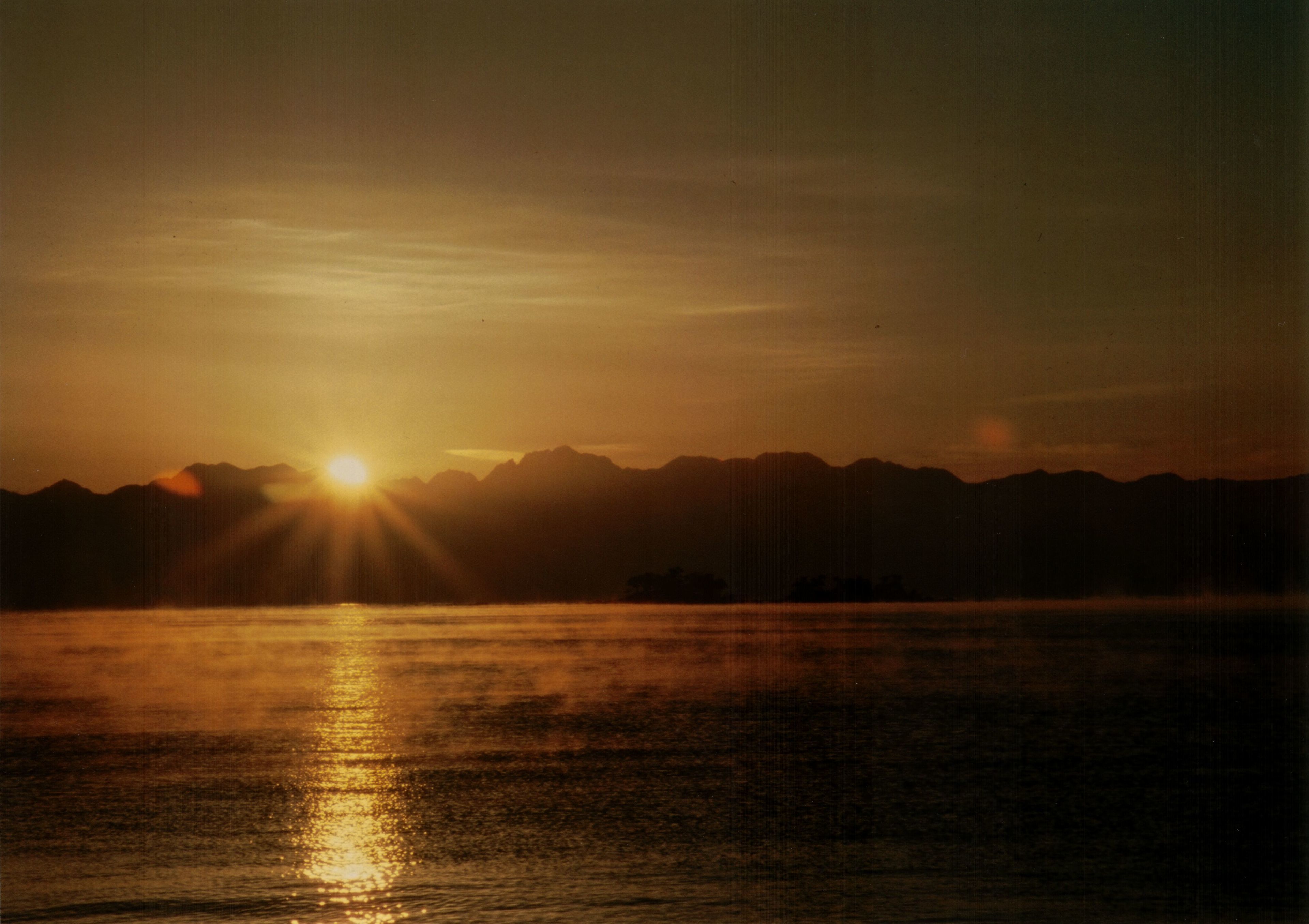 Ein Sonnenuntergang über einem Gewässer, im Hintergrund Berge.