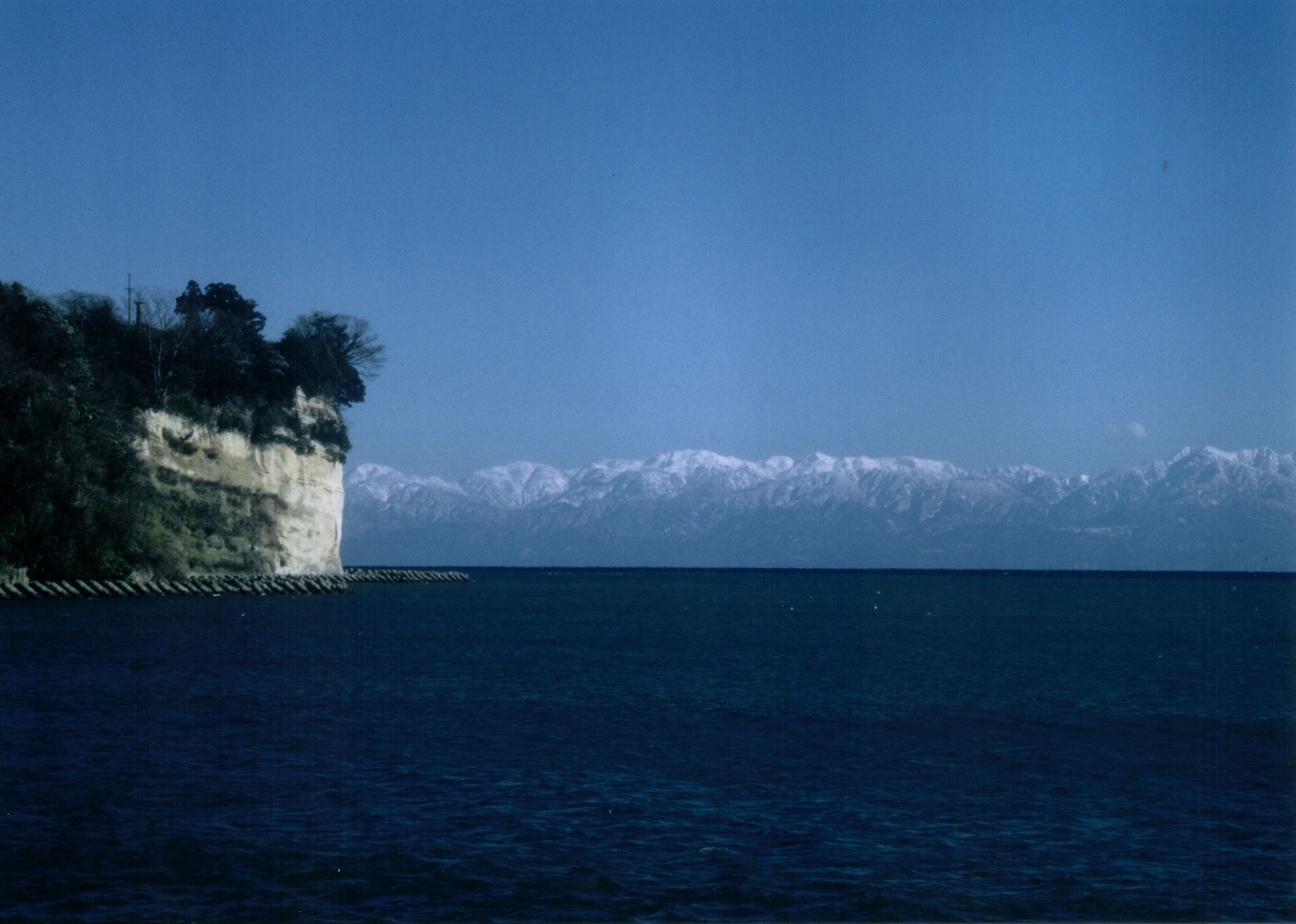 A coastal landscape with a cliff, a body of water, and snow-capped mountains in the background.