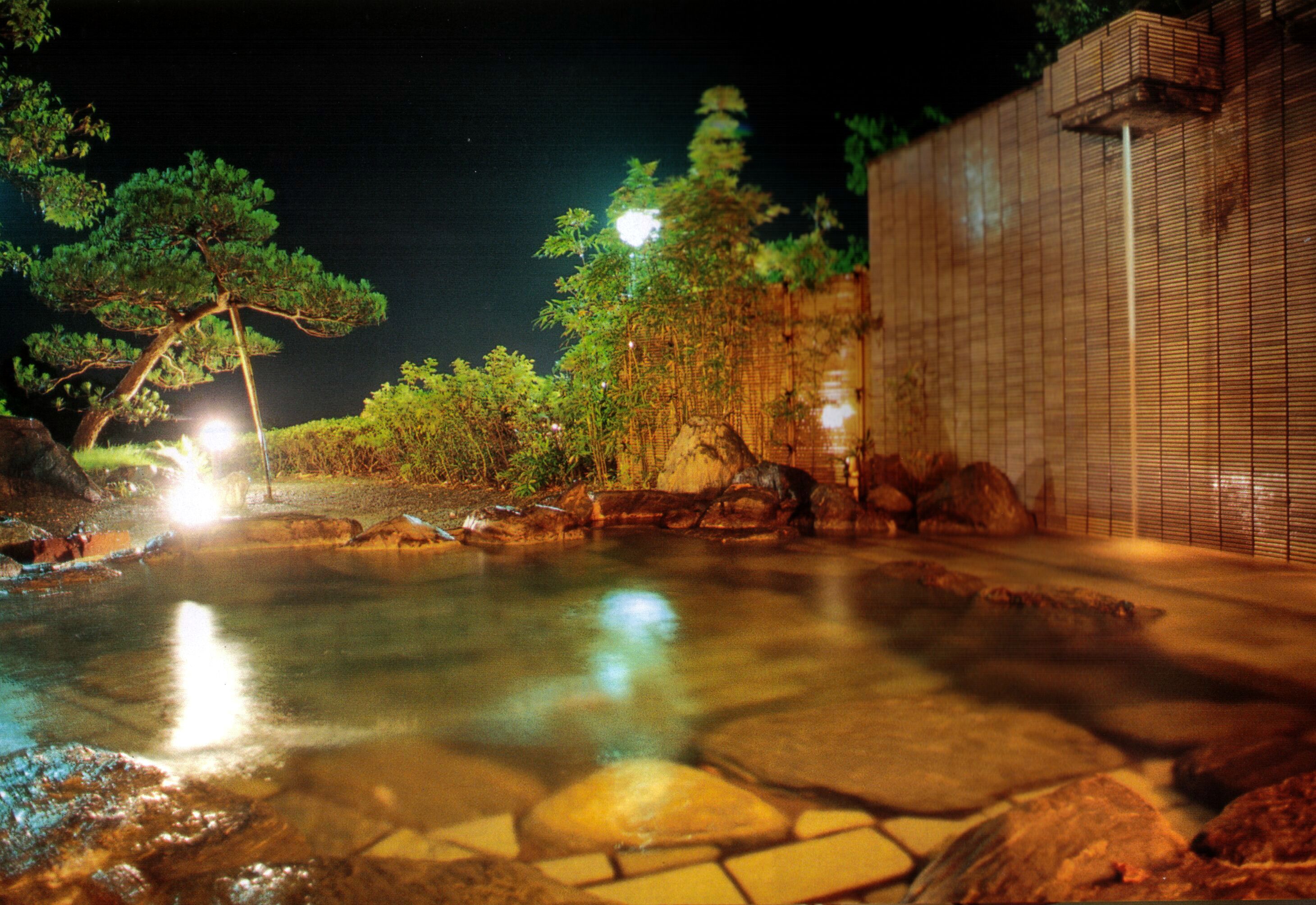 A nighttime view of a tranquil outdoor pool area with stone tiles, surrounded by trees and a bamboo fence.