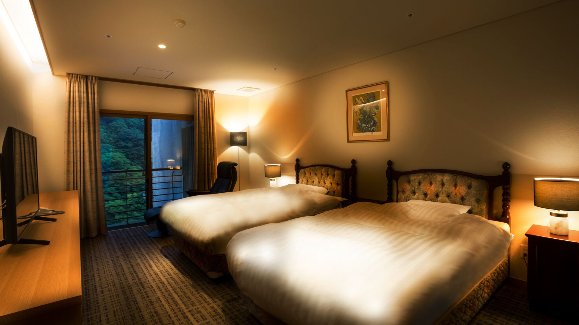 A hotel room with two beds, a desk, a chair, a TV, and a balcony with a view of greenery.