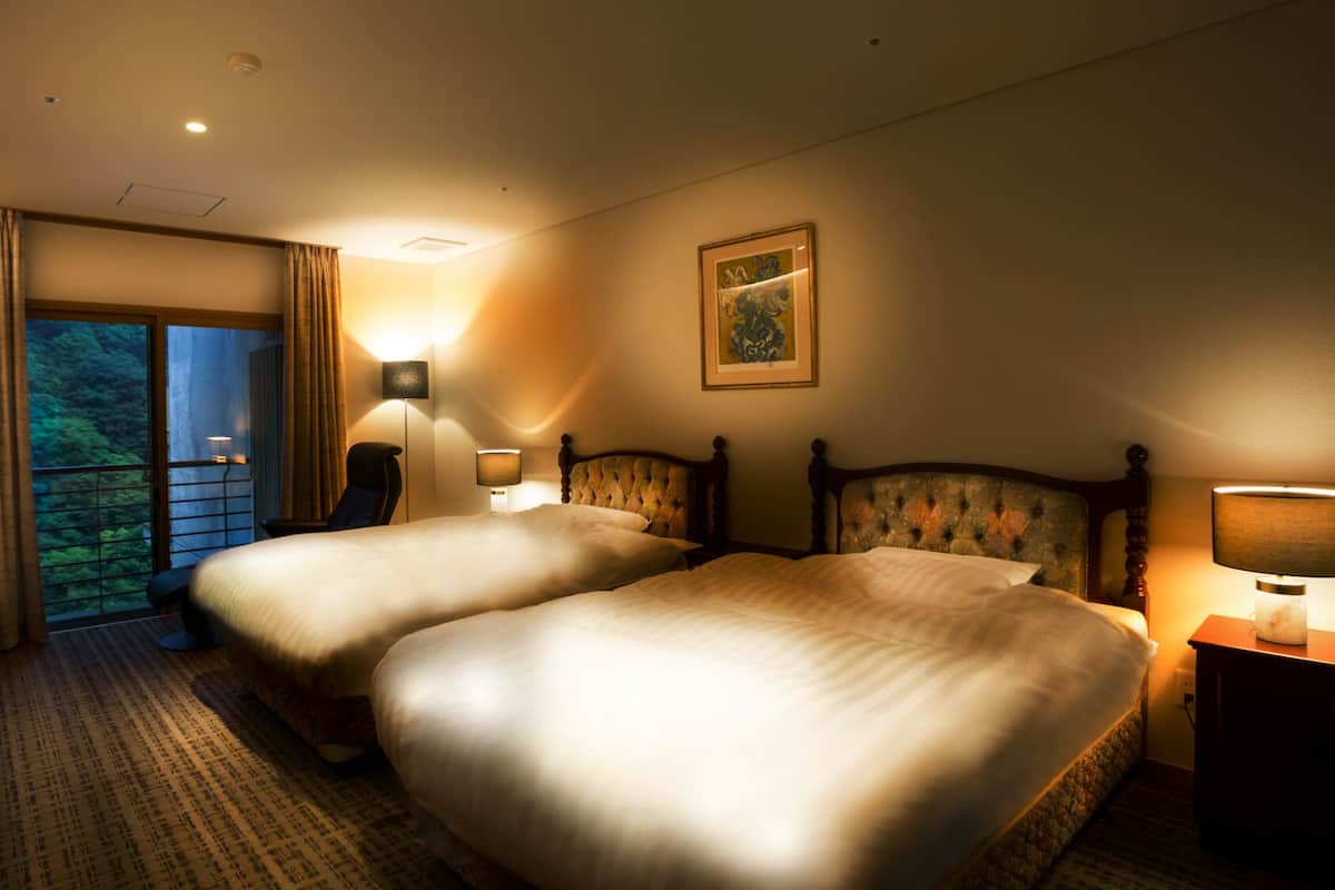 A hotel room with two beds, a desk, a chair, a TV, and a balcony with a view of greenery.