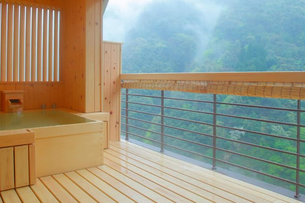 A wooden room with a wooden bathtub, a window with a view of a forest, and a bamboo blind.