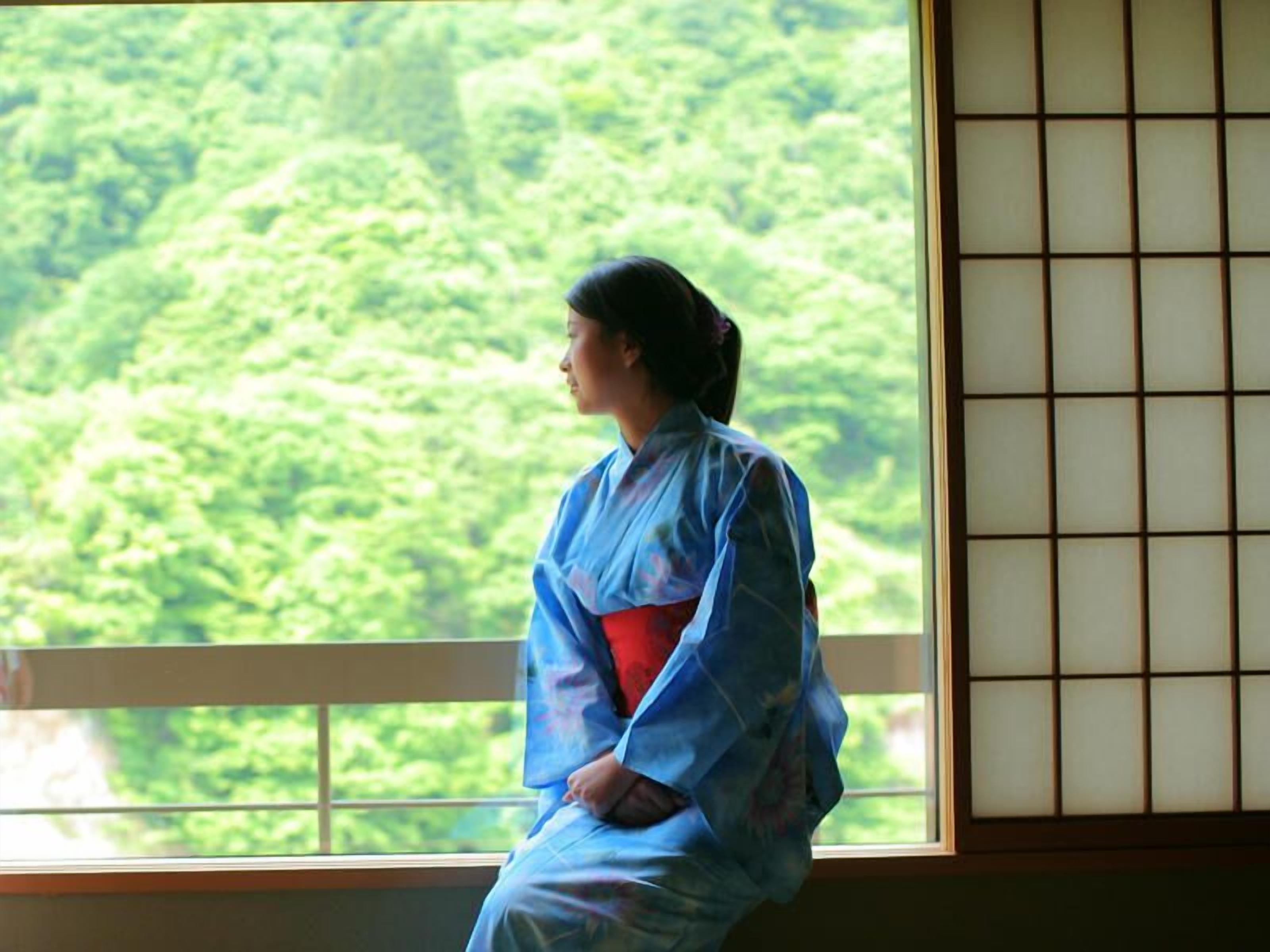 A person in traditional attire sitting by a window with a view of a forest.