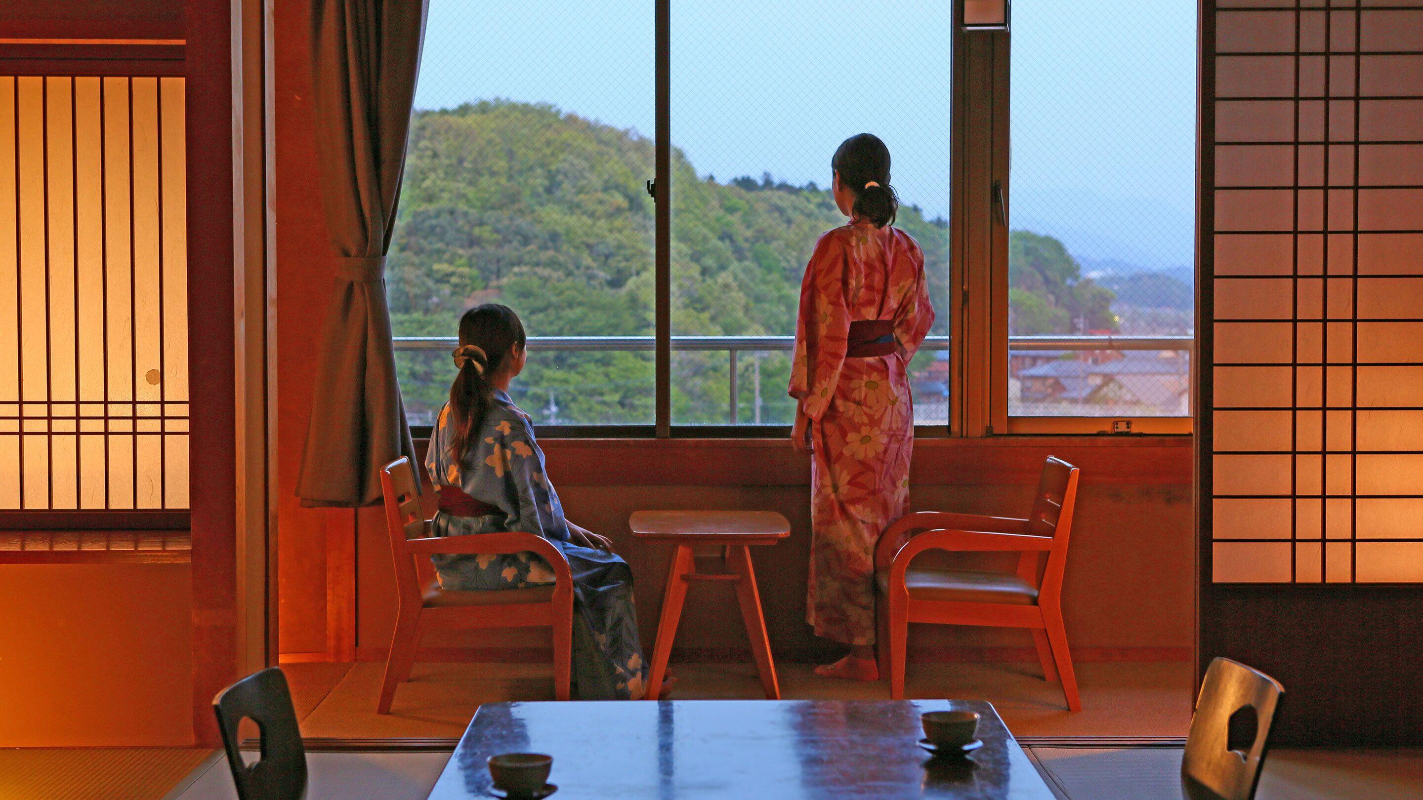 A traditional Japanese room with tatami flooring, a low table with a tea set, and a view of a hillside.