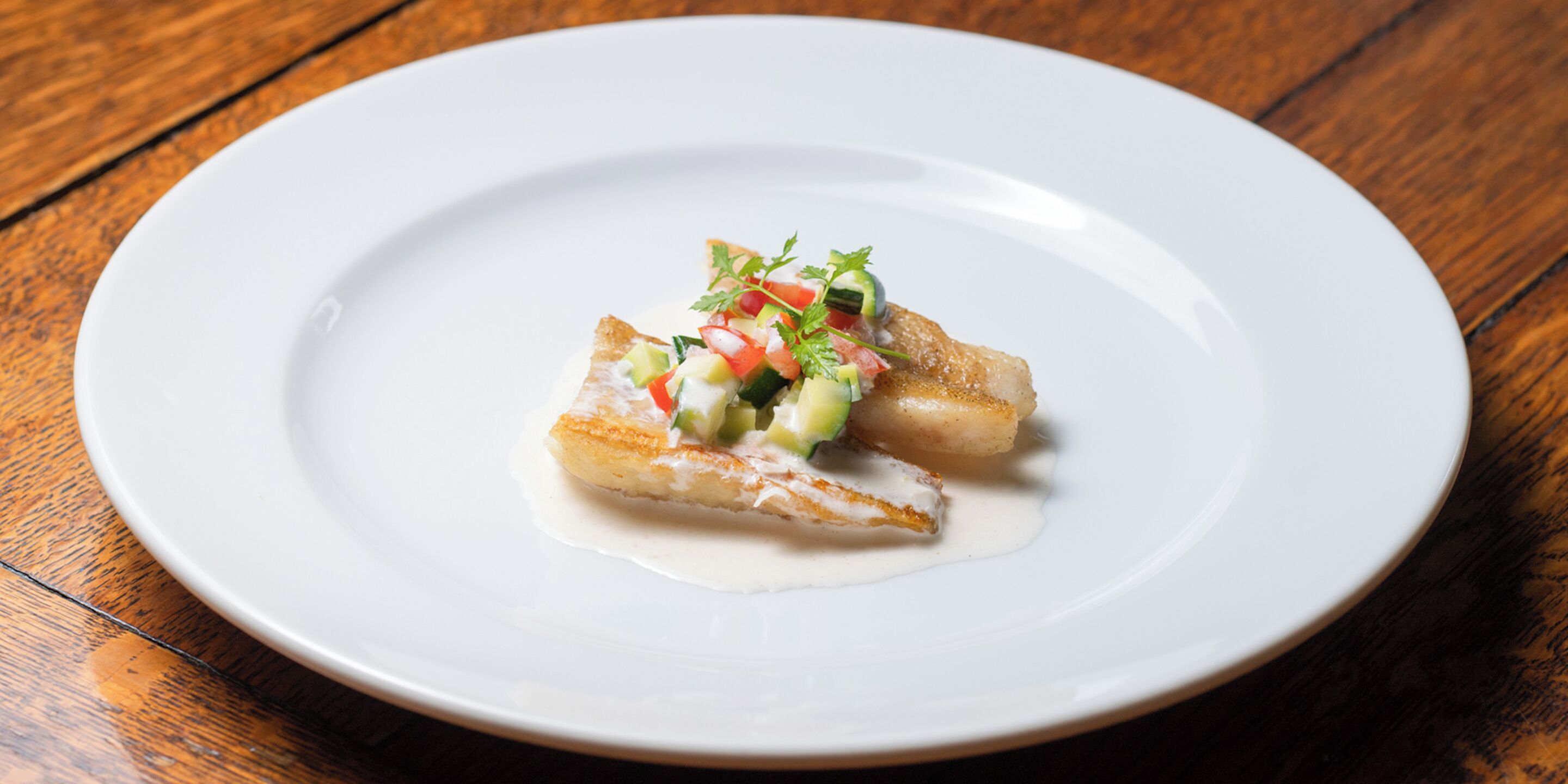 A white plate with a piece of fish and a small salad on a wooden table.