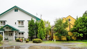 A two-story building with a green roof and white walls, surrounded by lush greenery and a small pond. - Gasthof Ami (Nikko)