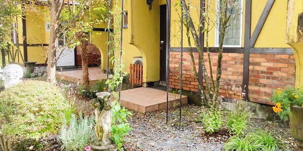 Un edificio amarillo con base de ladrillo y un pequeño jardín con una estatua y plantas.