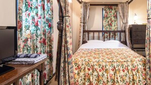 A bedroom with a wooden bed, floral curtains, a bedside table with a lamp, and a TV on a stand. - Gasthof Ami (Nikko)