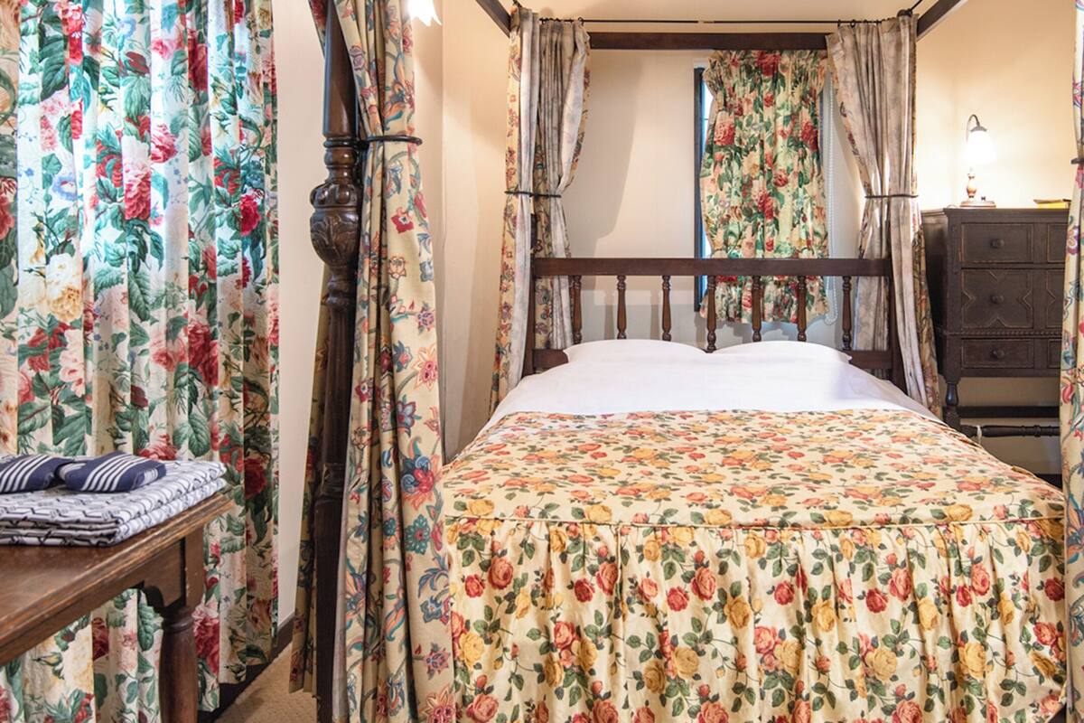 Un dormitorio con cama de madera, cortinas florales, una mesita de noche con lámpara y un televisor sobre un soporte.