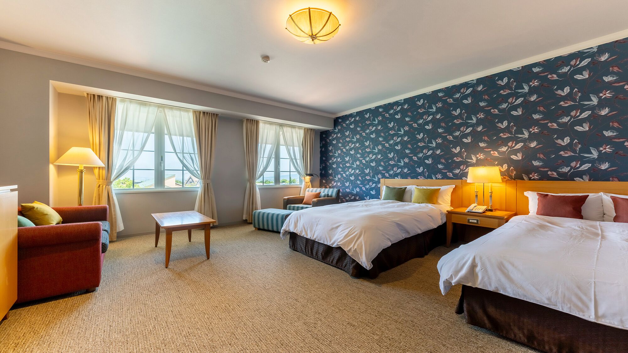 A hotel room with two beds, a small table, a chair, and a sofa. There are curtains on the windows, and the room has a carpeted floor.