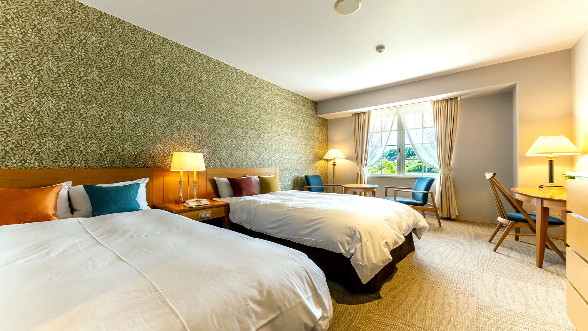 A hotel room with two beds, a desk, chairs, a window with curtains, and a patterned wall.