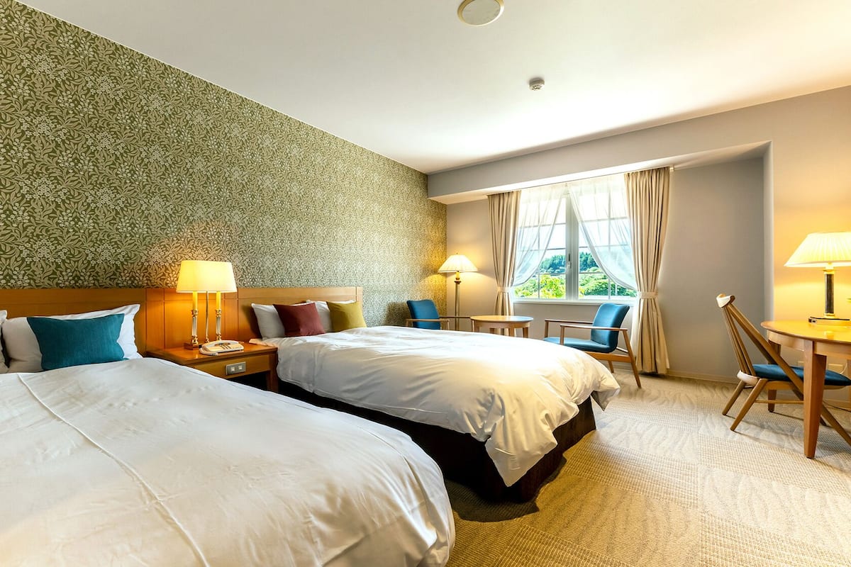 Habitación de hotel con dos camas, un escritorio, sillas, una ventana con cortinas y un papel tapiz estampado.