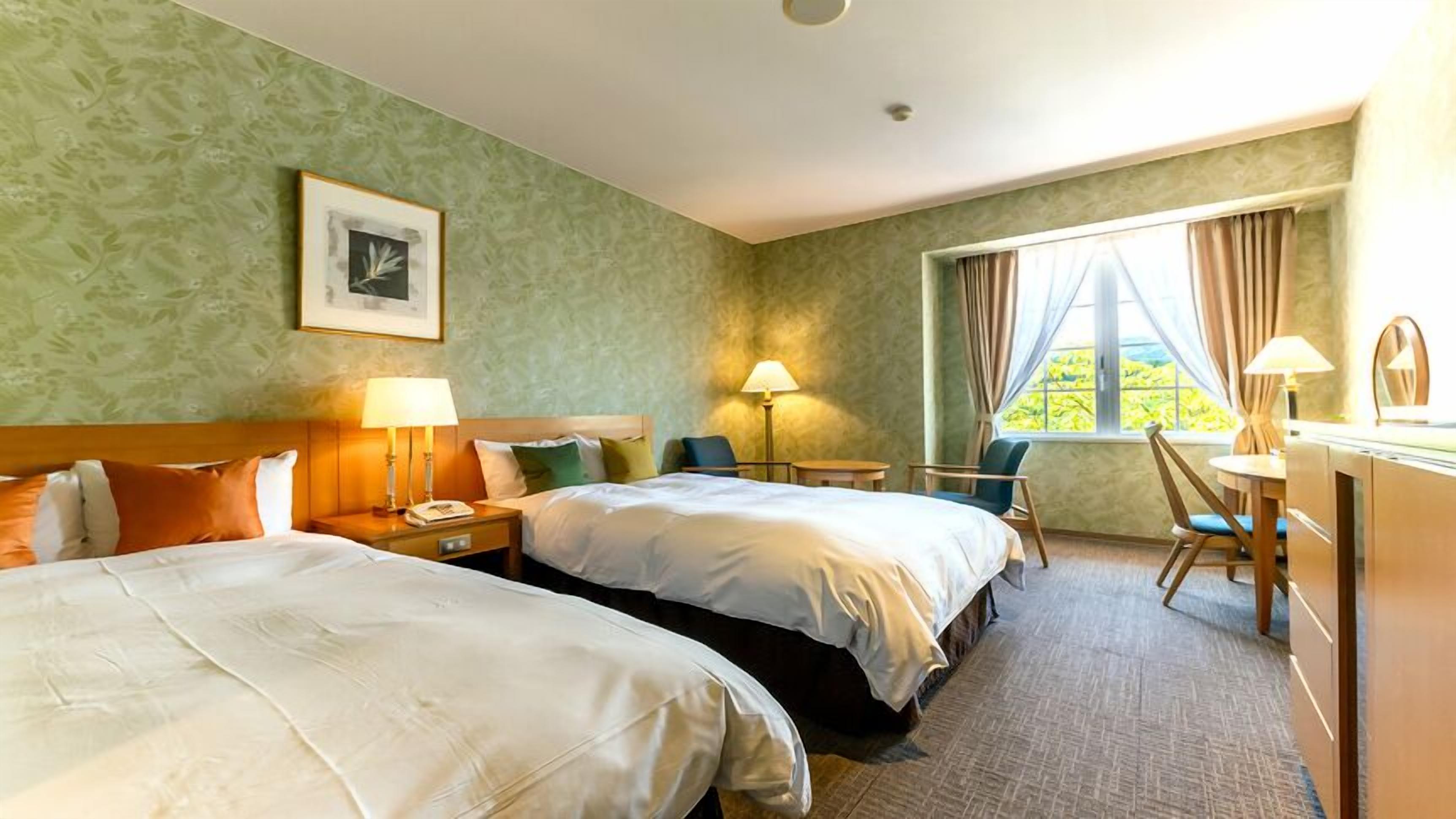 A hotel room with two beds, a desk, a chair, a lamp, and a window with curtains.