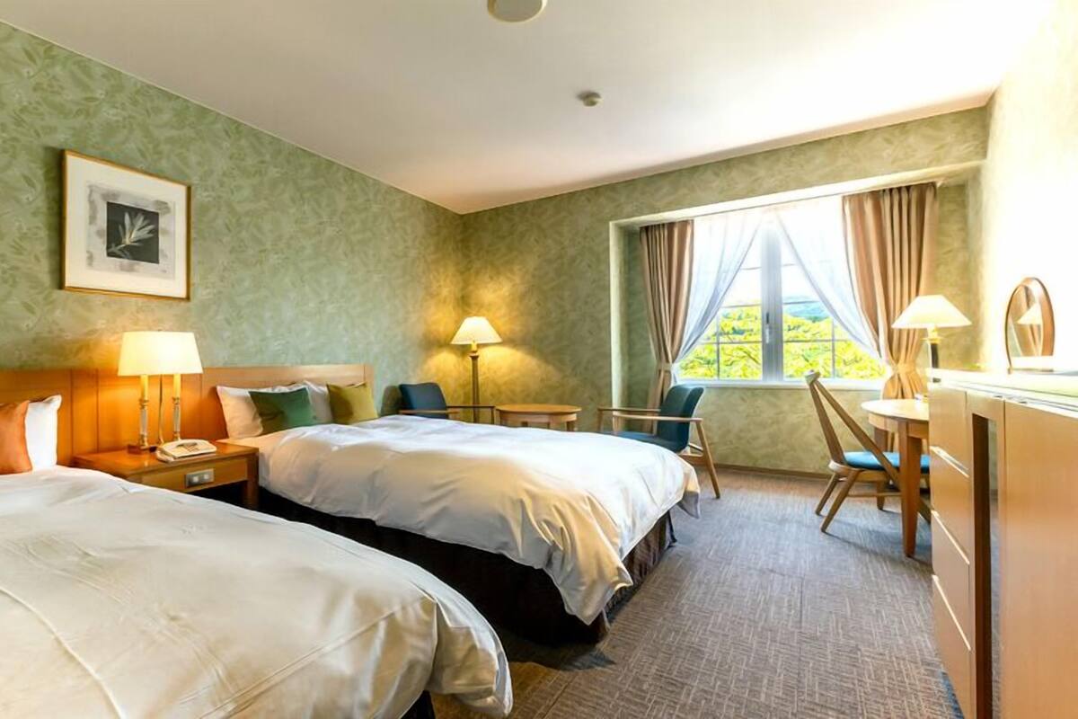 Habitación de hotel con dos camas, un escritorio, una silla, una lámpara y una ventana con cortinas.