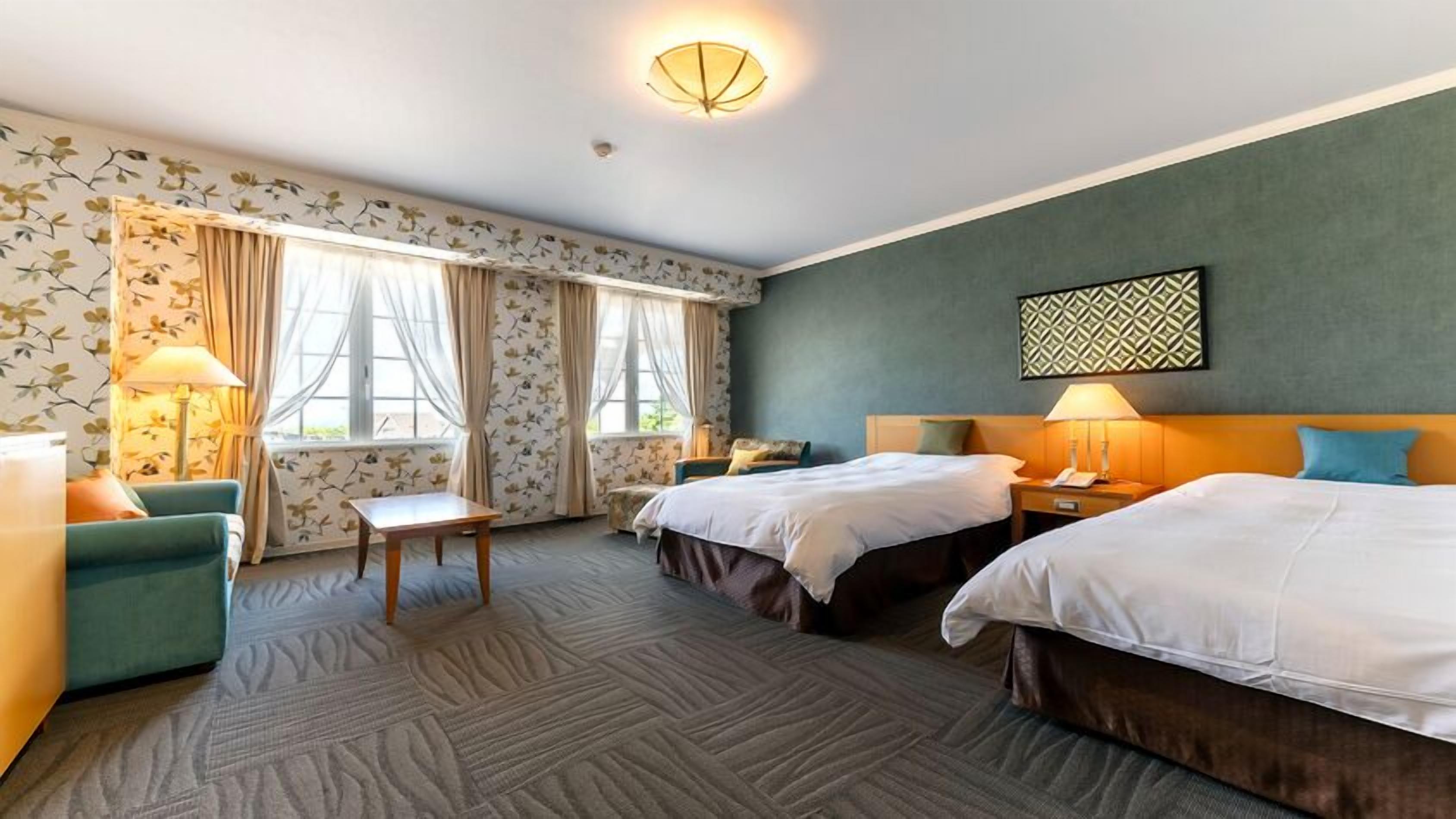 A hotel room with two beds, a desk, a chair, a lamp, and a window with curtains.