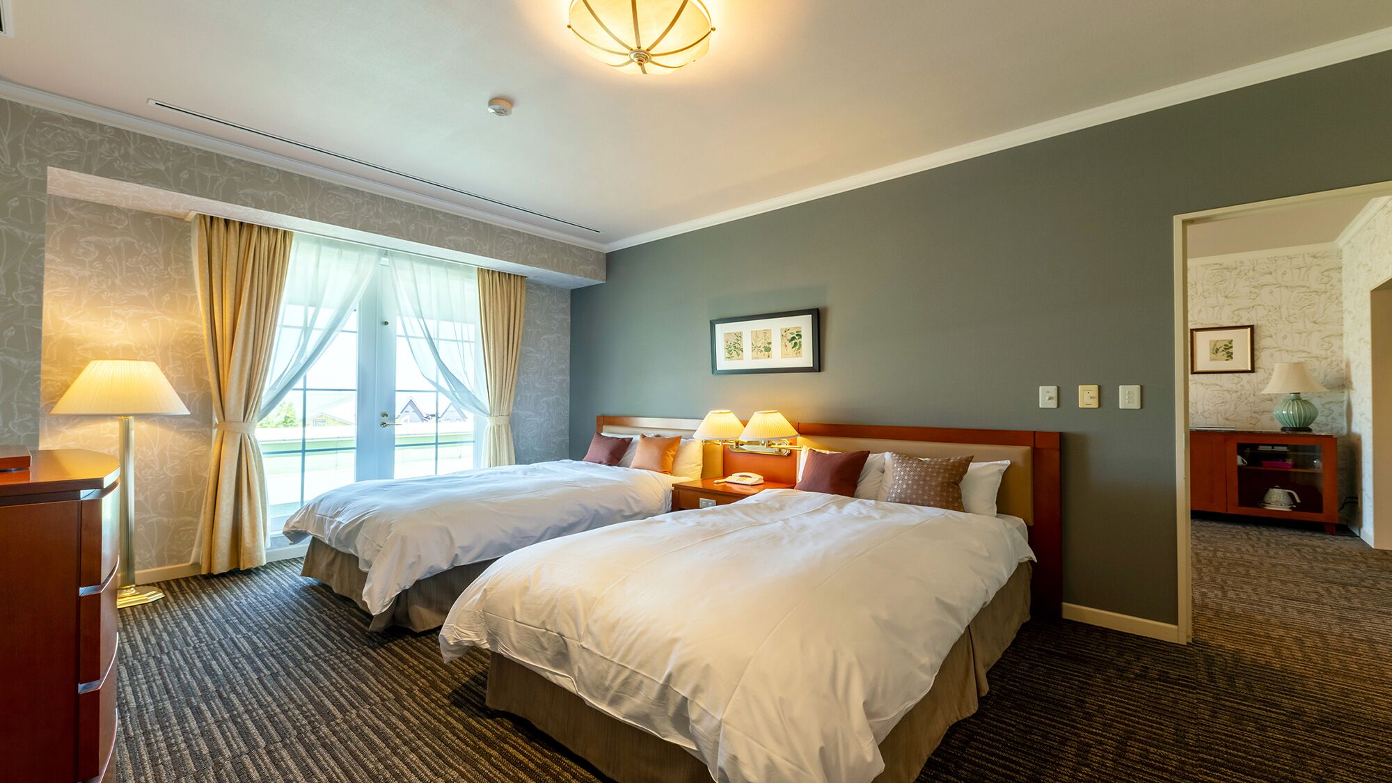 A hotel room with two beds, a nightstand with lamps, a door leading to another room, and a window with curtains.