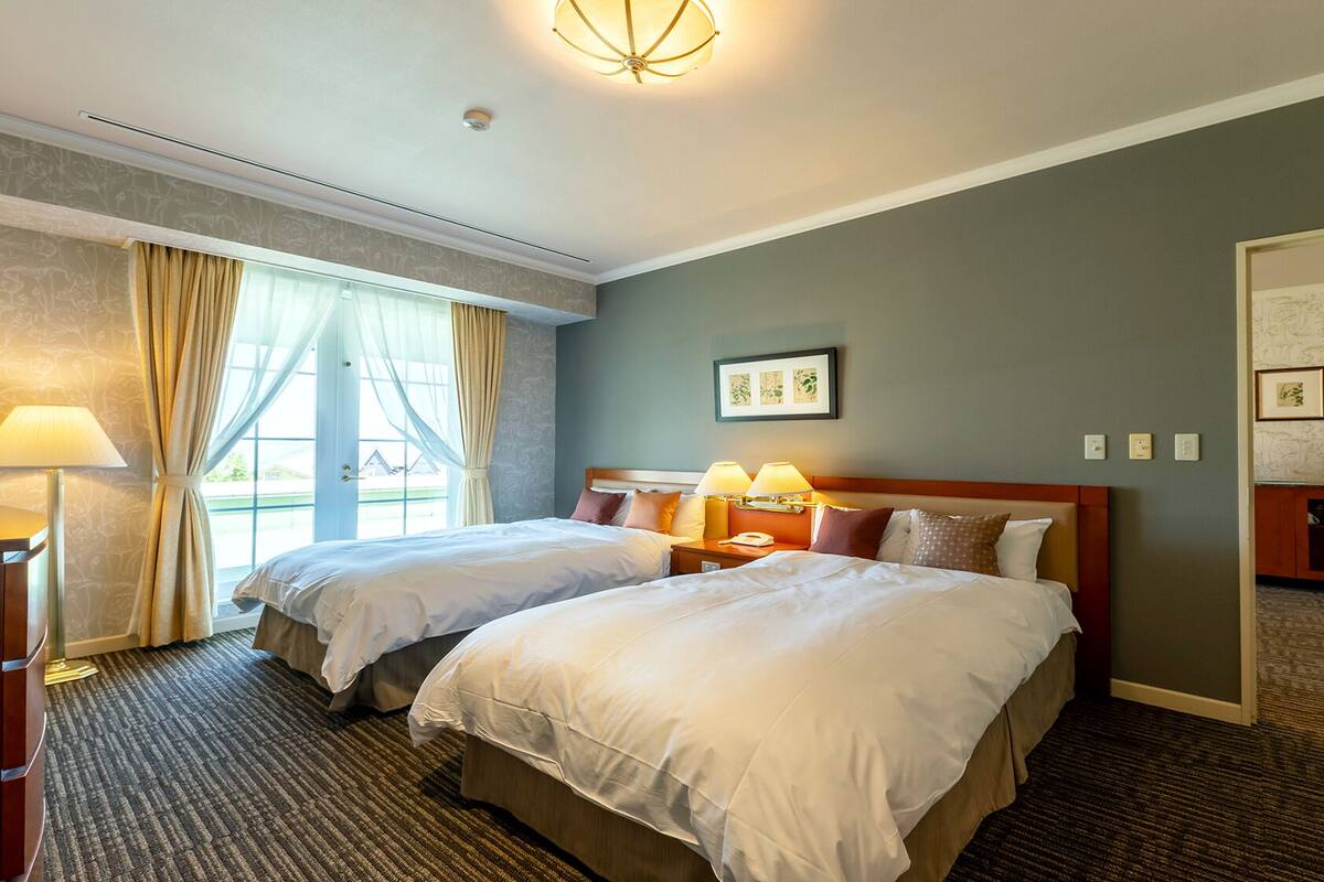 Habitación de hotel con dos camas, una mesita de noche con lámparas, una puerta que da a otra habitación y una ventana con cortinas.