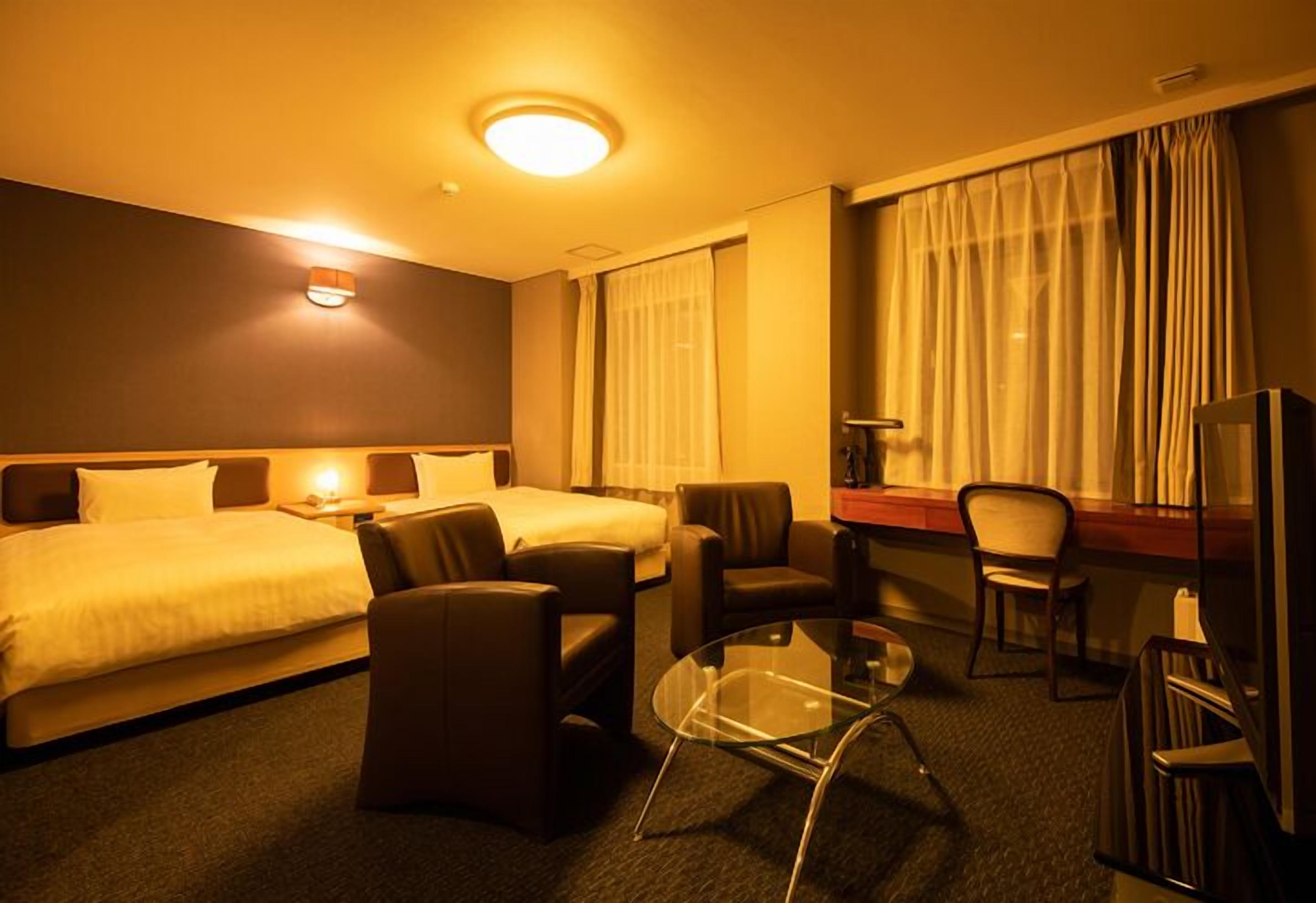 A hotel room with a bed, two armchairs, a glass coffee table, a TV, and a desk with a lamp.