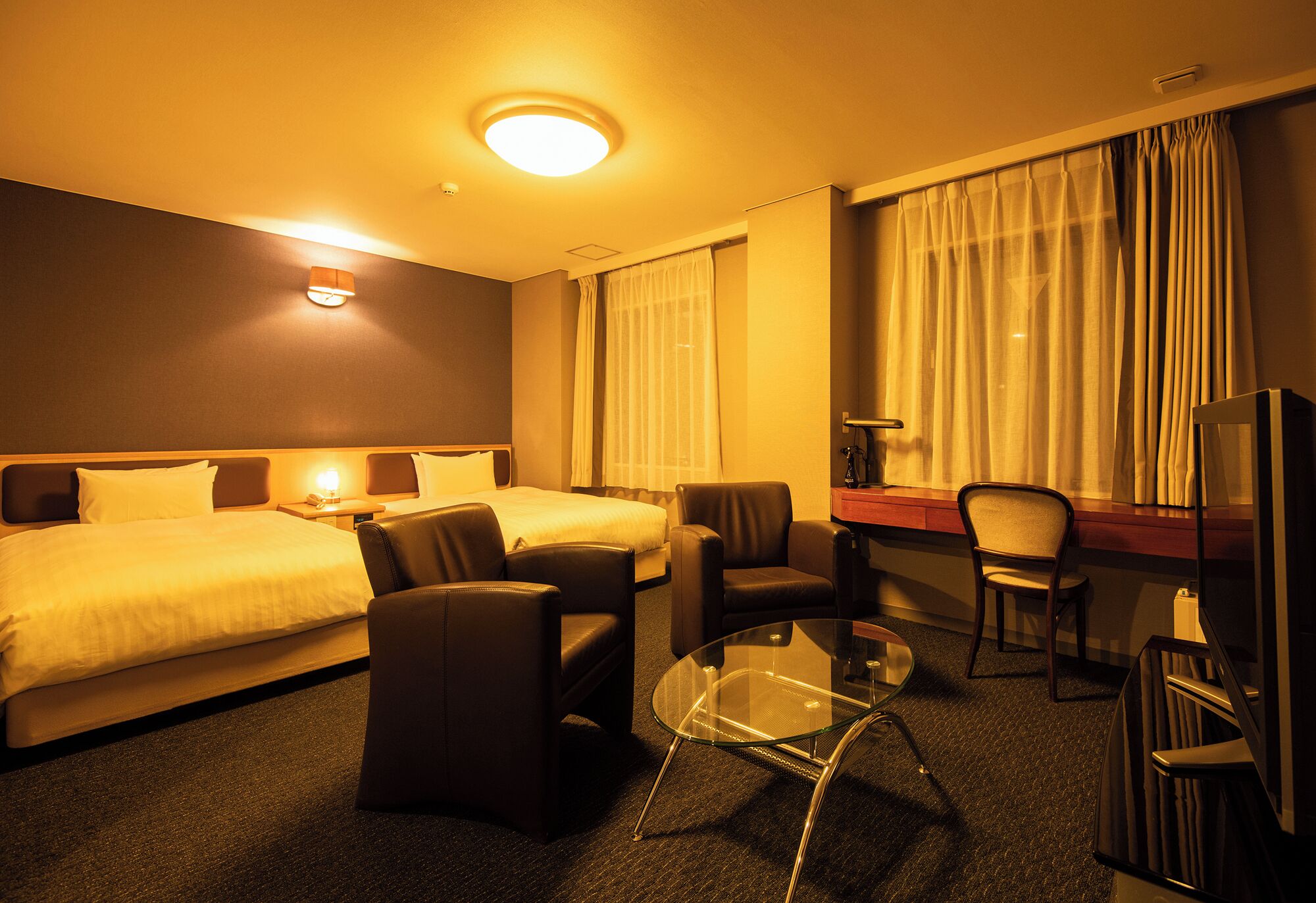A hotel room with a bed, two armchairs, a glass coffee table, a desk, and a television.