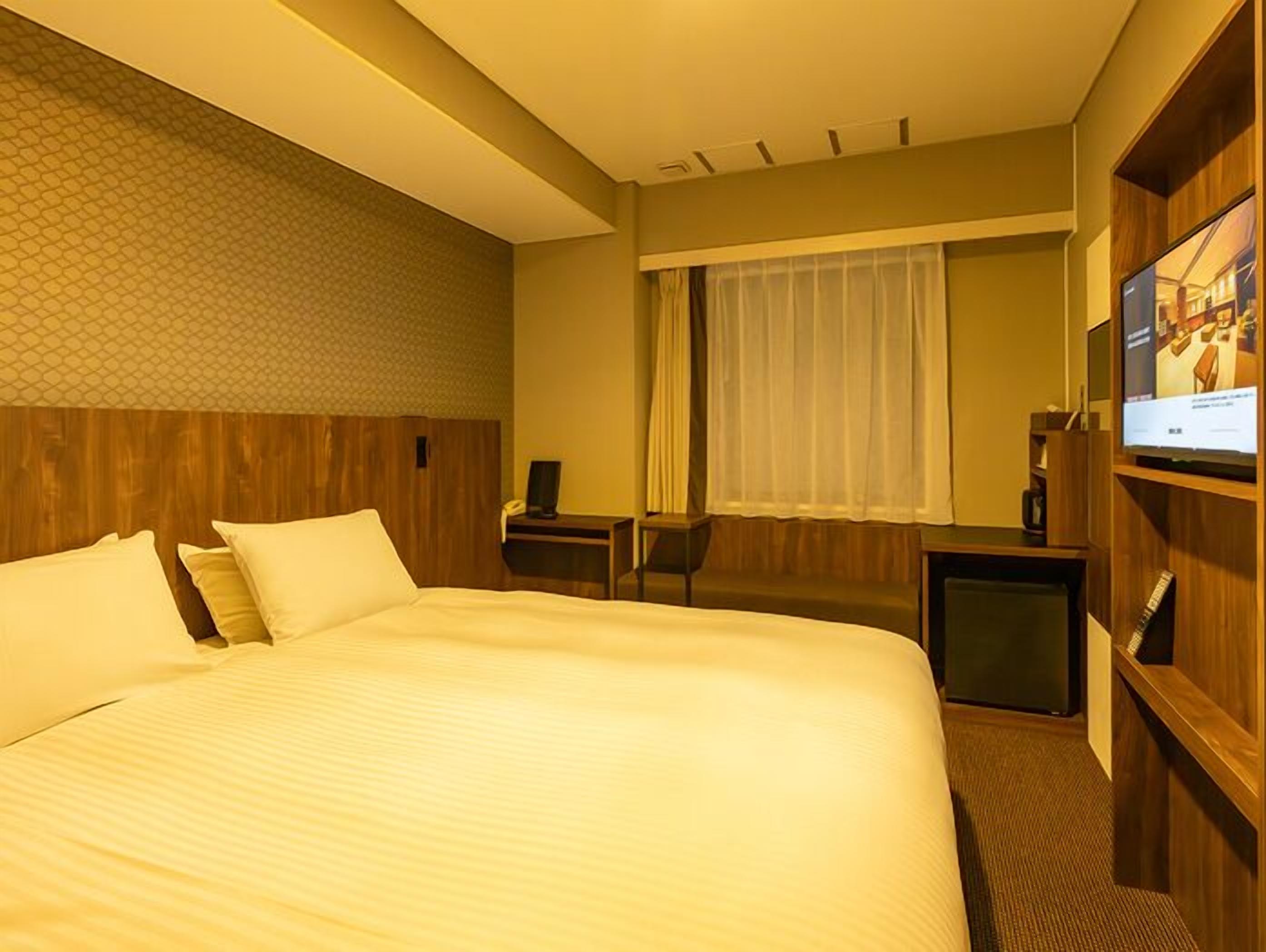 A hotel room with a large bed, a desk with a computer, a TV, and a window with curtains.