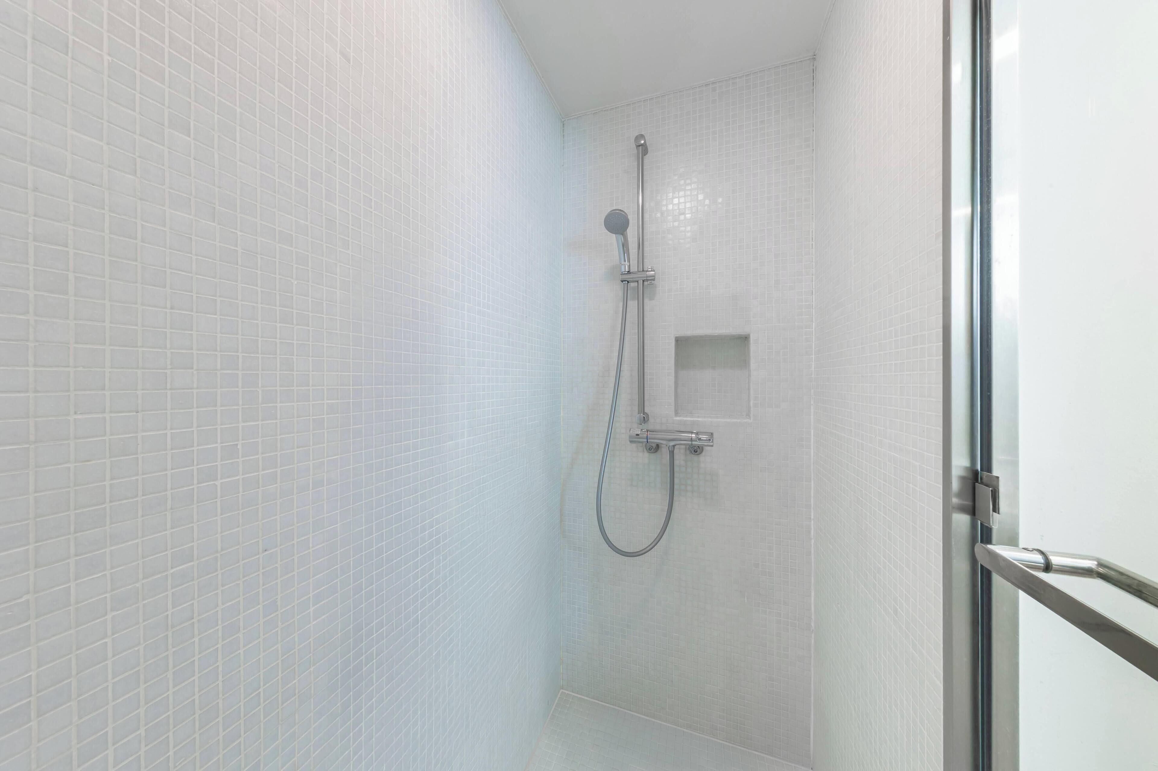 A bathroom with white tiled walls, a shower with a handheld showerhead and a handheld faucet, and a small square mirror.