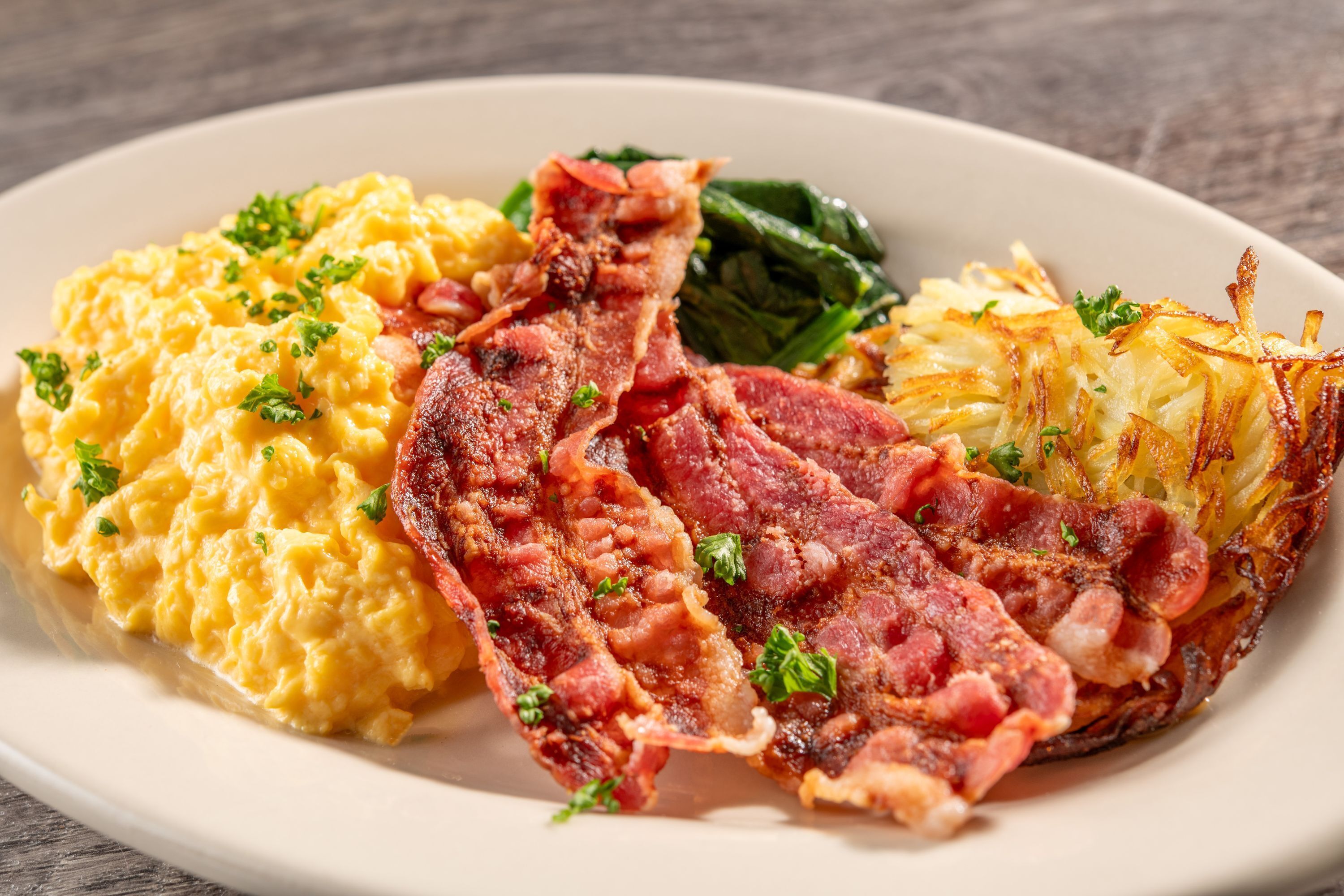 Un plato con huevos revueltos, tocino, papas salteadas y espinacas.