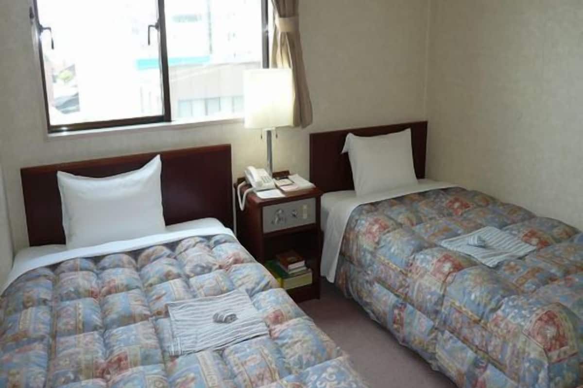 A hotel room with two beds, a nightstand with a lamp, a window with curtains, and a view of the outside.