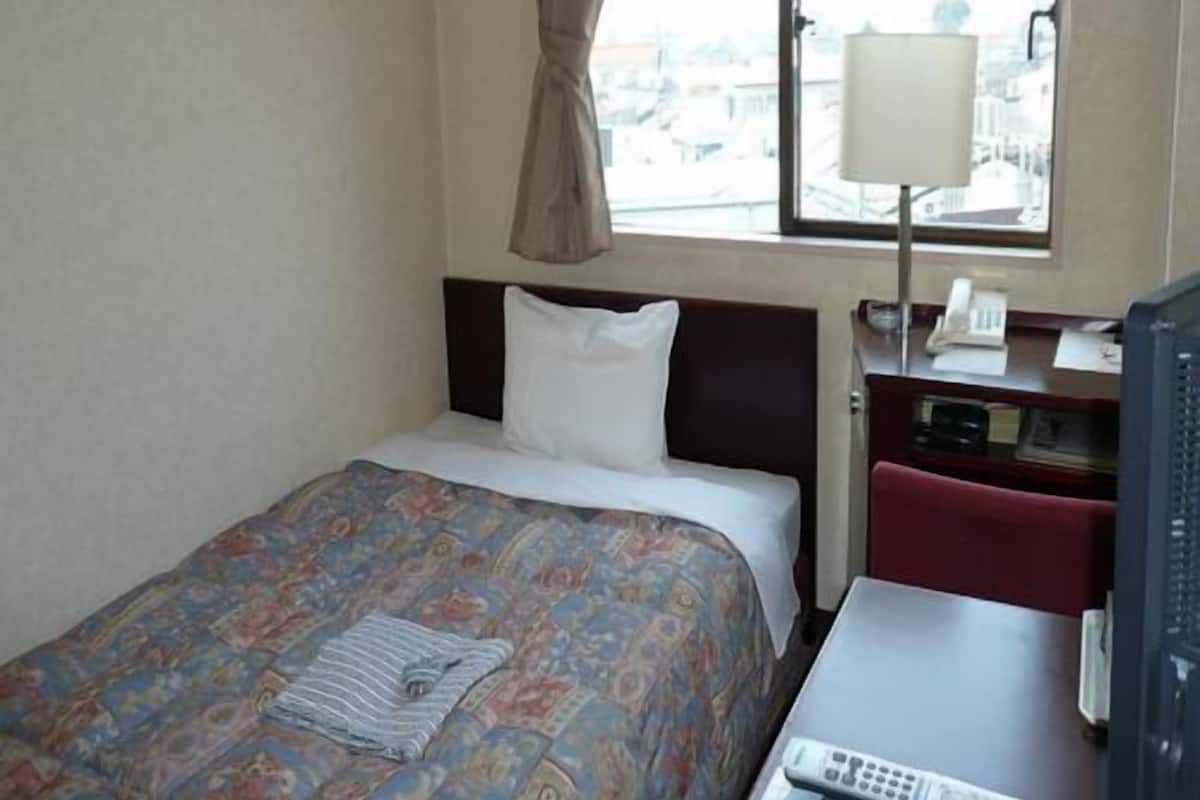 A hotel room with a bed, a desk with a telephone and a remote control, a window with a curtain, and a view of buildings outside.