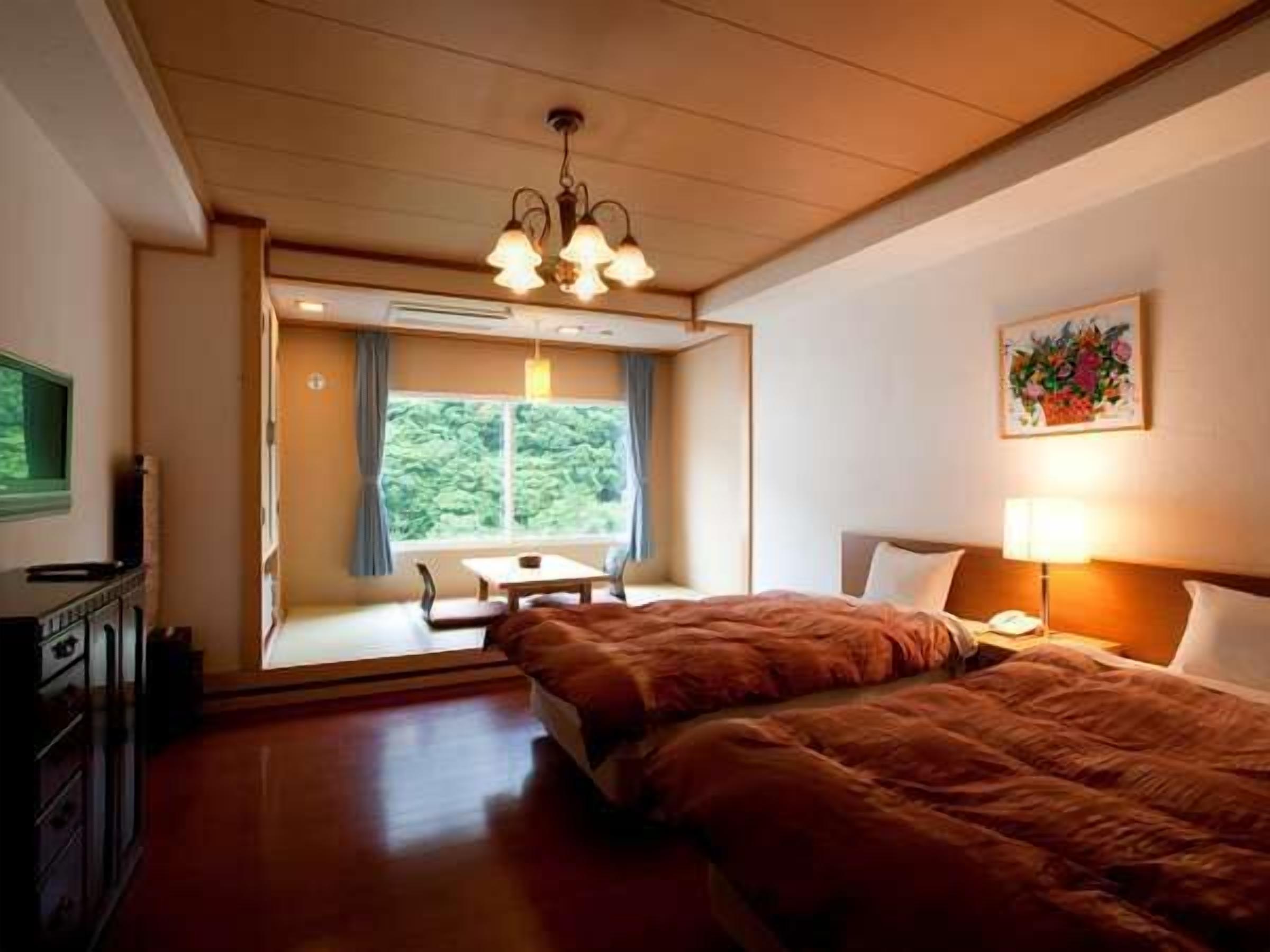 Ein Hotelzimmer mit Bett, Nachttisch, Lampe und einem Fenster mit Blick auf Grünflächen.