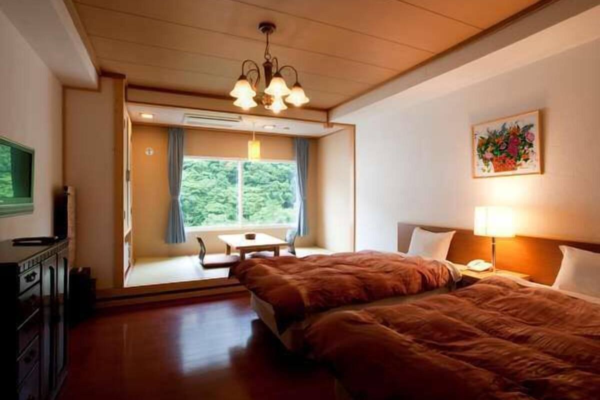 A hotel room with a bed, bedside table, lamp, and a window with a view of greenery.