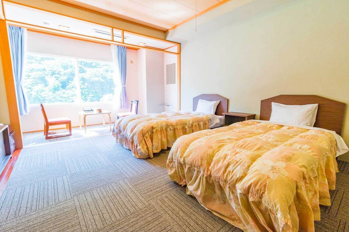 A hotel room with two beds, a chair, a small table, and a view of trees through the window.