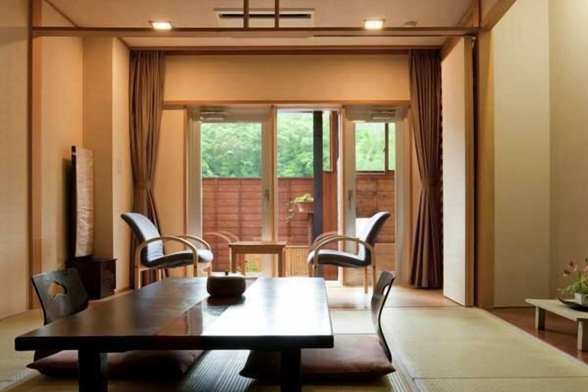 A room with a wooden table, four chairs, a tatami mat floor, and a view of greenery outside.