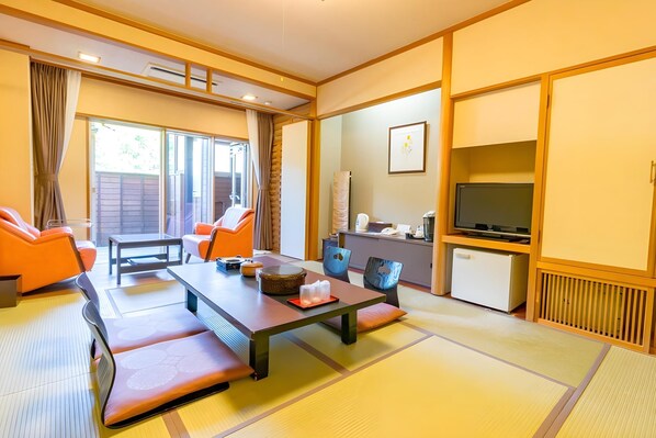 A traditional Japanese-style room with tatami flooring, a low table, orange chairs, a television, and a sliding door leading to a balcony.