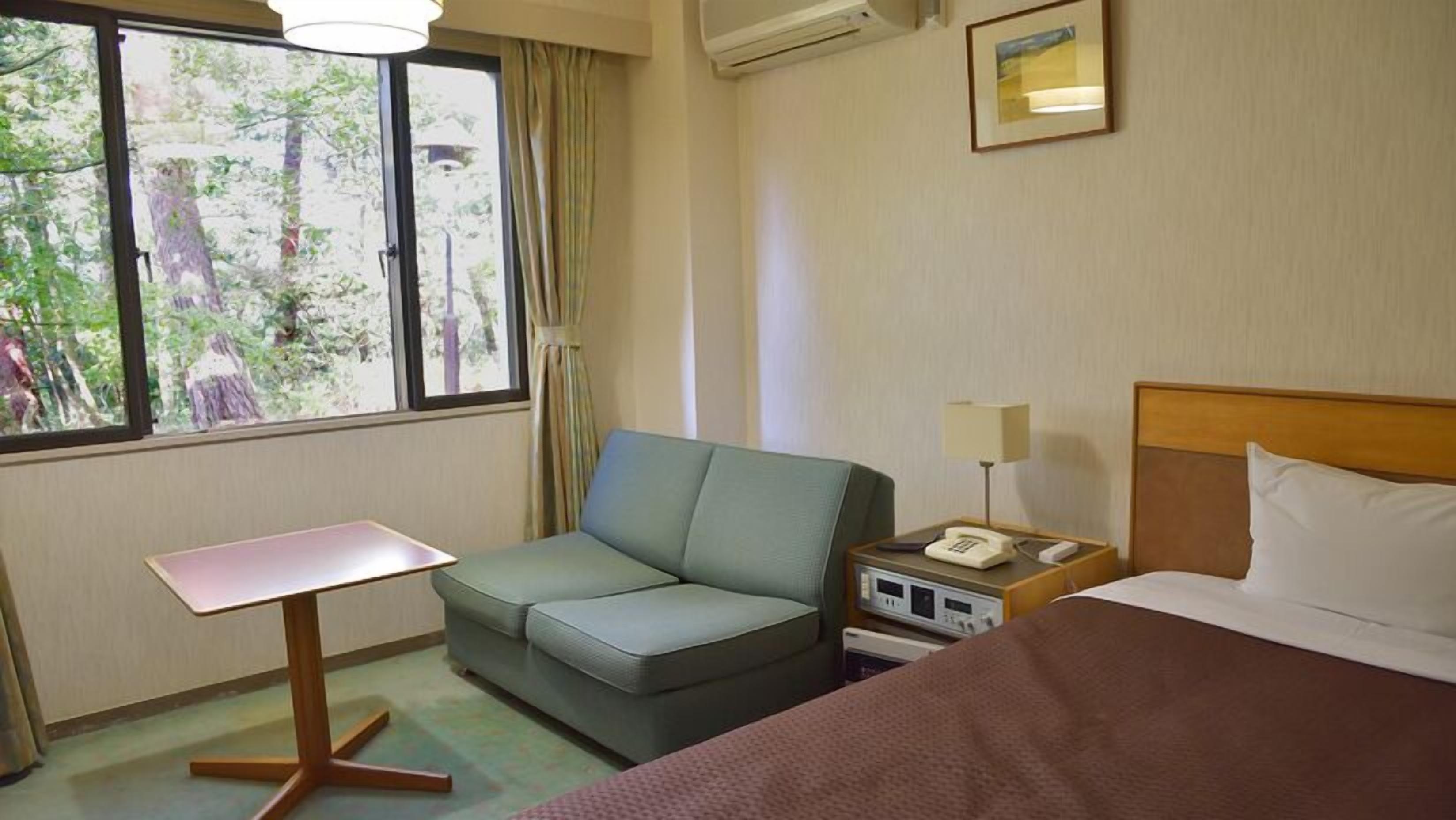 A hotel room with a bed, a sofa, a small table, a nightstand with a lamp, and a window with a view of trees.