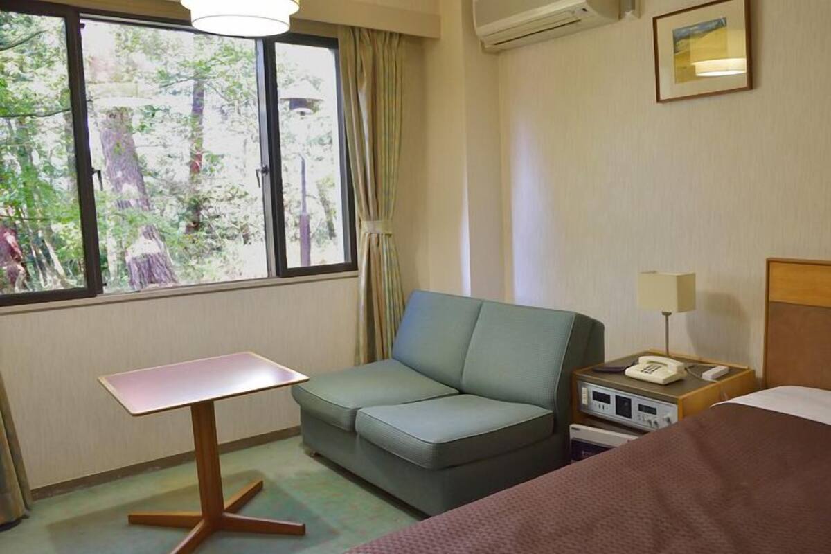 A hotel room with a bed, a sofa, a small table, a nightstand with a lamp, and a window with a view of trees.