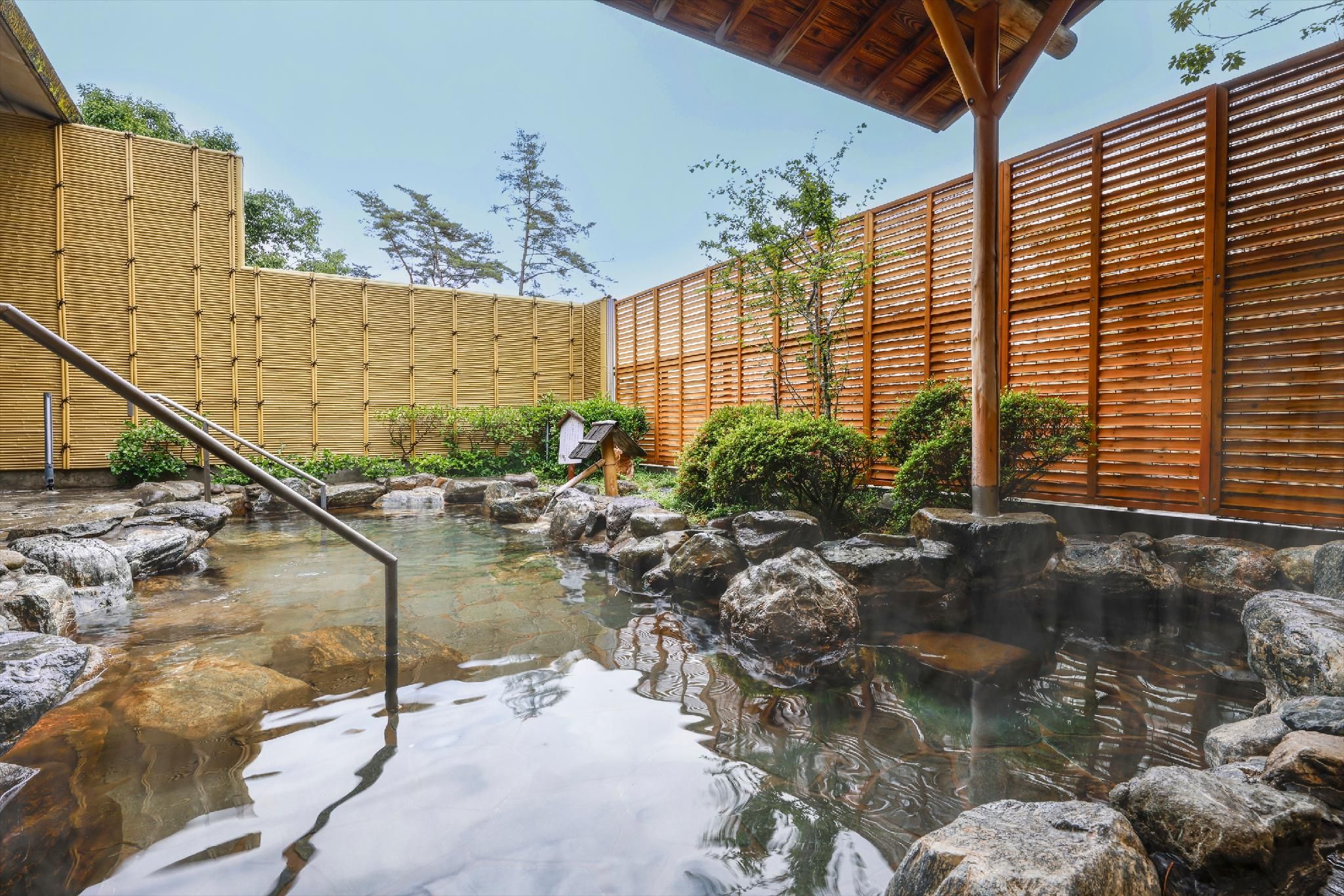 A traditional Japanese onsen with a wooden fence, a stone hot spring, and a wooden walkway.