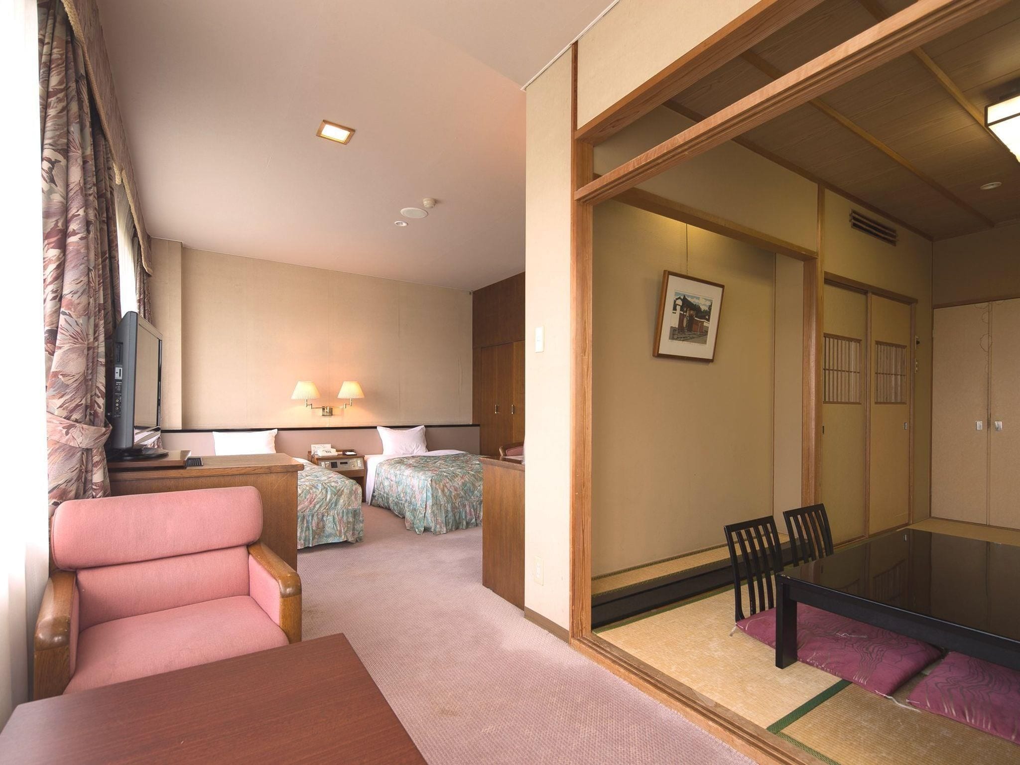 A hotel room with a bed, two bedside tables, a desk, a chair, a dining table, and a sliding door leading to another room.