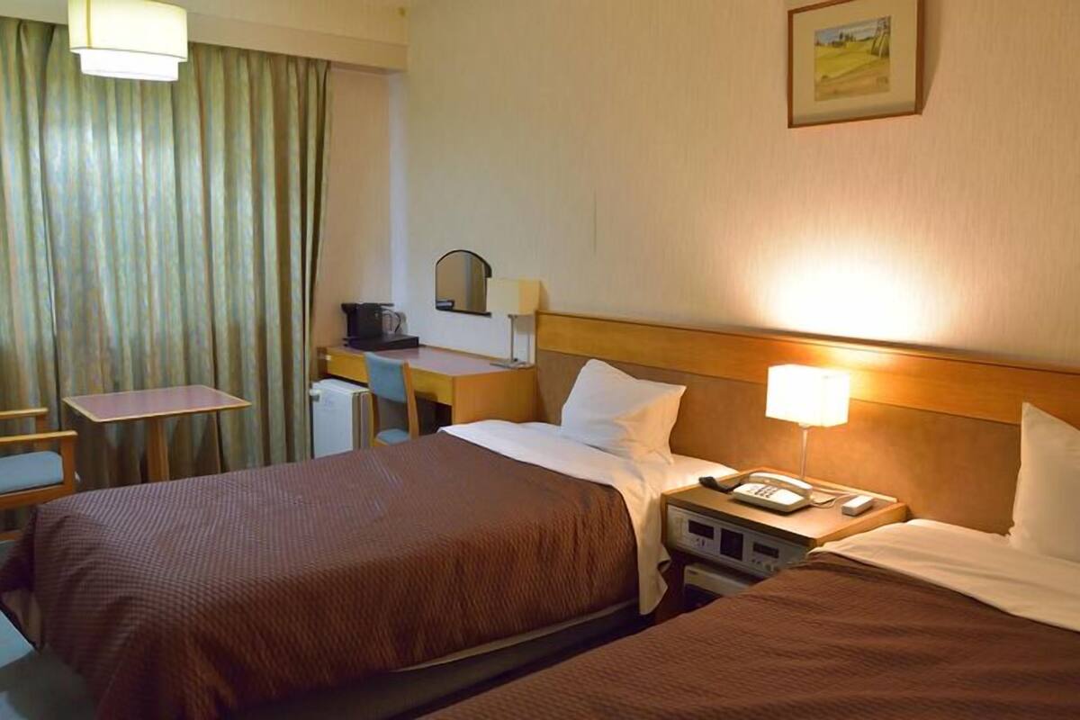A hotel room with two beds, a desk, a chair, a telephone, and a painting on the wall.